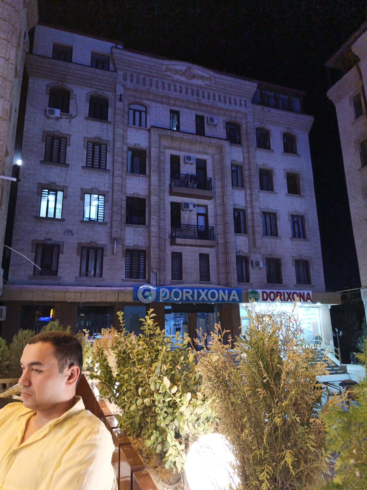 In an outdoor nighttime scene, a man in a yellow shirt sits to the left of the frame, looking to his left. Behind him, a multi-story building with a lit facade stands tall. The building features numerous windows, some with visible interior lights. A sign reading "DORIYONA" is visible on the lower part of the building, indicating a pharmacy. Various green shrubs and plants are in the foreground, partially obscuring the view of the building's lower section. The overall lighting suggests a dimly lit urban environment.