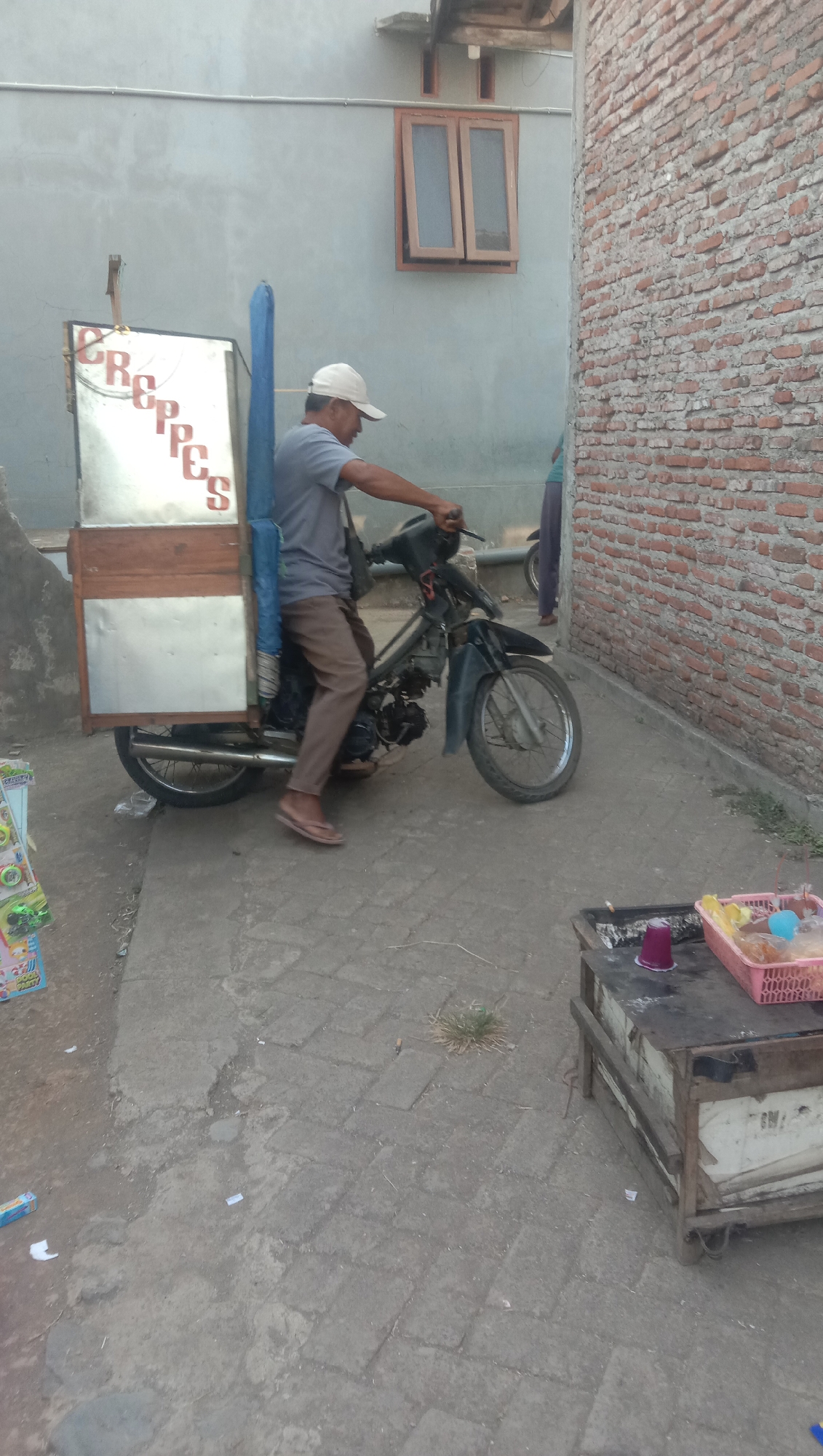A man wearing a grey t-shirt, brown pants, and a white baseball cap is riding a motorcycle with a food cart attached. The food cart has a silver and wooden exterior and is labeled with the word "CREPES". The man is positioned on the motorcycle, facing right, with his hands on the handlebars. He is wearing flip-flops. The motorcycle is dark-colored, with a visible engine and wheels. The background shows a grey building with a window and a brick wall. To the left of the man, there's a stack of colorful items, possibly merchandise or packaging. In the foreground, to the right, is a wooden cart or table with items on it, including a pink basket containing what appear to be food items or snacks. The ground is paved with interlocking grey stones, some of which are broken or uneven, with patches of dirt and weeds. The scene appears to be an outdoor street or alleyway in a city.