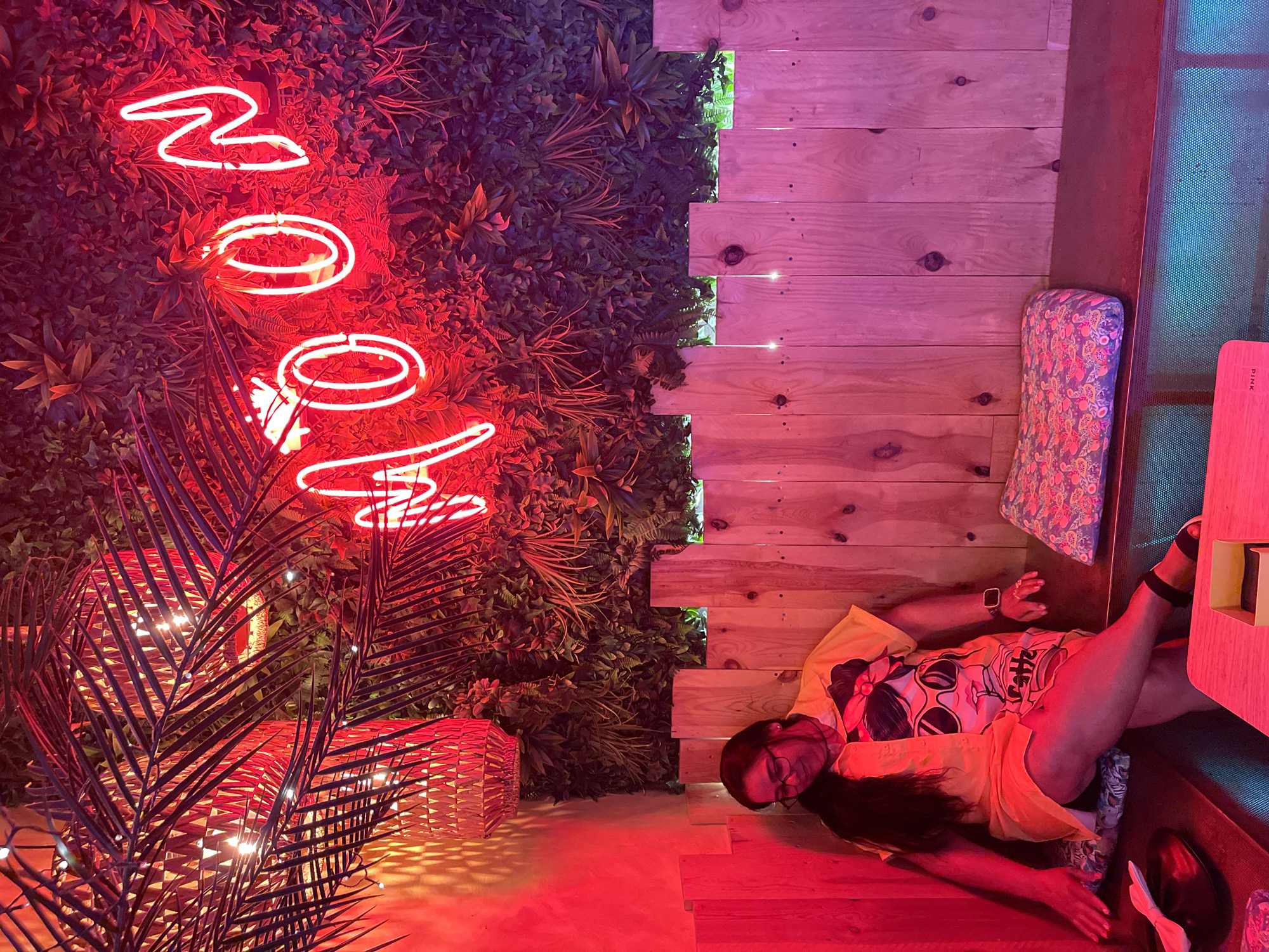 A woman sits on a bench in a dimly lit establishment. The walls are adorned with artificial foliage and illuminated by a bright pink neon sign spelling "MOON." She wears a yellow shirt with a graphic print and glasses, with her legs crossed. To her right, a patterned cushion rests on the bench. A wooden fence-like structure separates the seating area from the background. In the foreground, a black hat and a small yellow box are visible on a surface. The lighting is predominantly red and pink. The scene appears to be in Barcelona, Spain.
