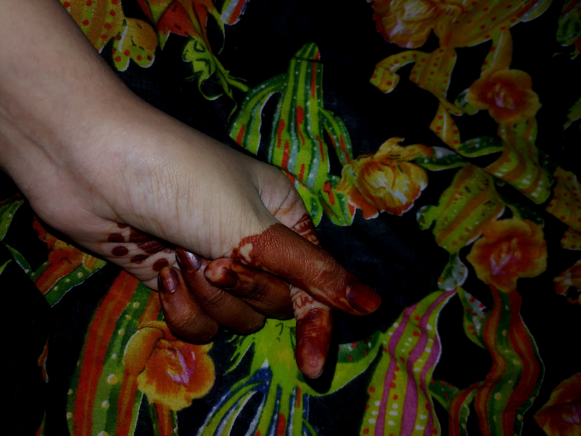 A left hand with henna designs on the fingers and palm rests on a black fabric with a vibrant floral print in yellow, orange, green, and red. The hand is lightly tanned, and the henna is a deep reddish-brown. The fabric has abstract floral and stripe patterns. The image is a close-up shot, focusing on the hand and the texture of the fabric. The background is the printed fabric. There is no information in the image to suggest a location in Thane, India.