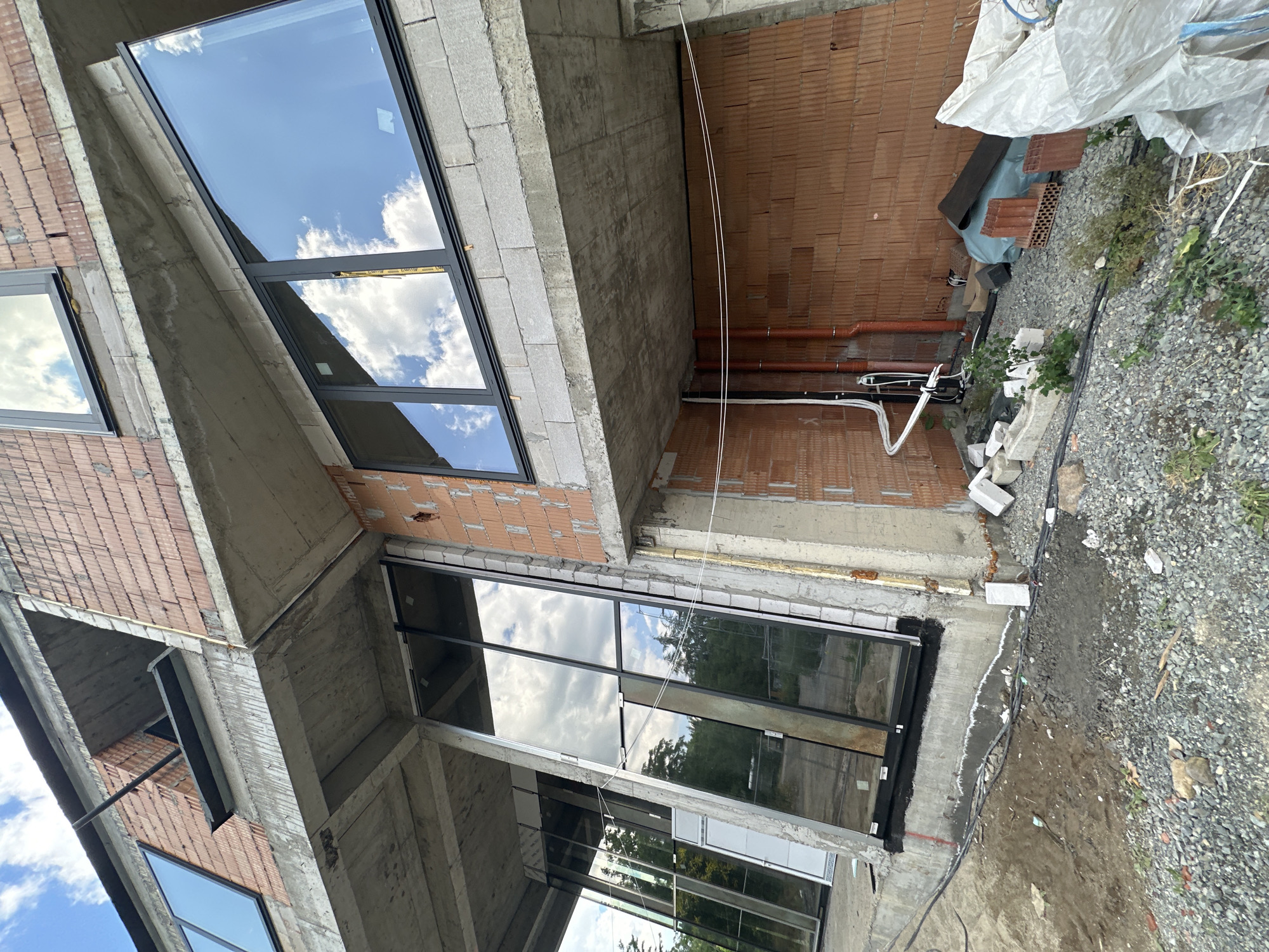 The image depicts an exterior view of a building under construction, possibly a multi-story residential or commercial structure, located in Stara Zagora, Bulgaria. The building's framework is predominantly concrete with exposed brick walls, indicating an ongoing construction phase. Several large windows with dark frames have been installed, reflecting the bright, cloudy sky.
The scene shows a construction site with various building materials scattered around, including bricks, gravel, and some white sheeting. Exposed plumbing pipes and electrical wiring are visible on the exterior walls. The ground around the building is uneven, with sections of dirt and gravel. The overall impression is one of active construction, with elements of the building's facade and surrounding site visible. There is no visible text in the image. The lighting suggests it is daytime, with clear skies and scattered clouds.
