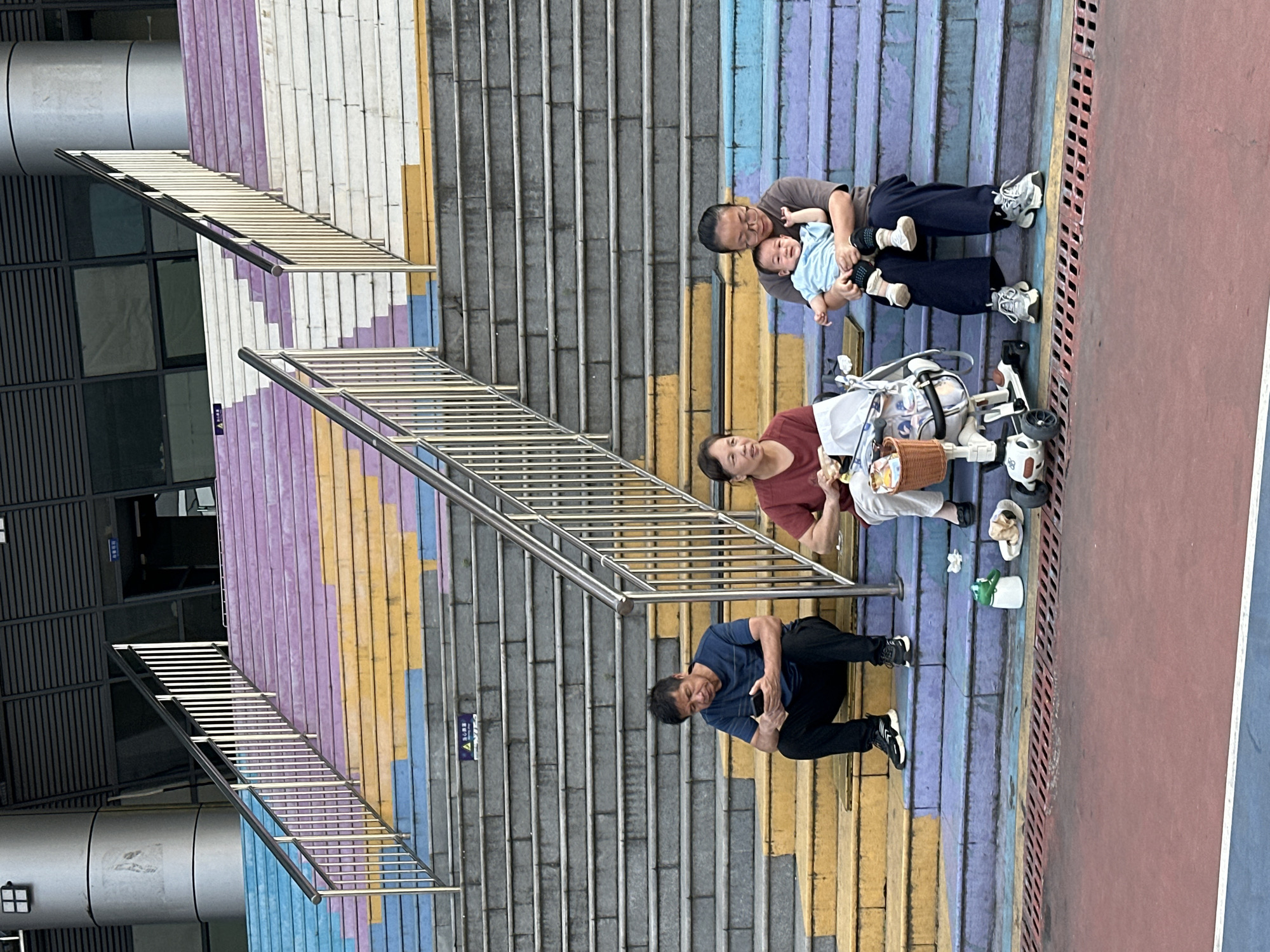 The image shows a family resting on a staircase in Banan, China. The staircase is painted with alternating colors of white, purple, yellow, and blue. A man is sitting on a lower step looking at his mobile phone. Above him, a woman sits next to a baby stroller. A man holding an infant sits further up on the stairs. Personal items, including shoes and what appears to be trash, are scattered on the steps next to the woman. Stainless steel handrails run alongside the steps. A building is visible behind the stairs.