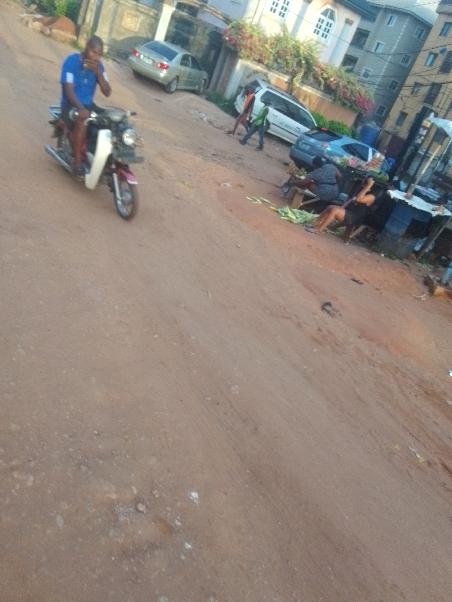 The image depicts a street in Nkpor, Nigeria. The road is unpaved and dusty. A man in a blue shirt is riding a motorbike. Several parked cars are visible, along with people on foot and two people sitting on a bench near some produce. There are buildings in the background.