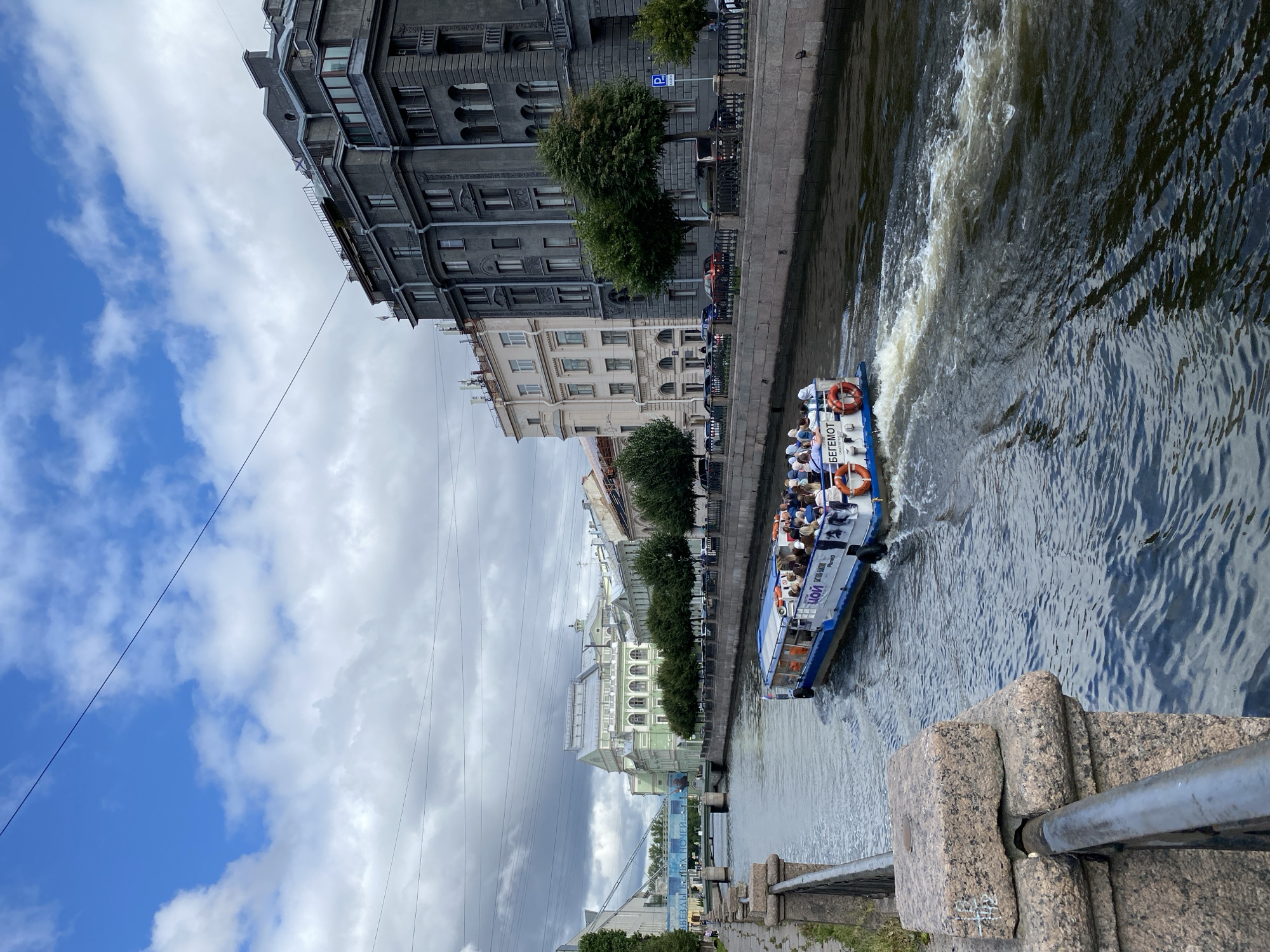 This is a vertical, eye-level shot of the Griboyedov Canal in Saint Petersburg, Russia, on a cloudy day. A tour boat named "BEGEMOT" is moving away from the viewer towards the left of the frame, with passengers visible on its upper deck. The canal is lined with stone embankments and trees. On the right bank, several multi-story buildings with classic architecture are visible. On the left bank, a walkway runs alongside the canal. In the background, a bridge spans the canal and another large, light-green building with a grey roof is prominent. The sky is a mix of blue and white clouds, with some power lines crisscrossing the upper portion of the image.