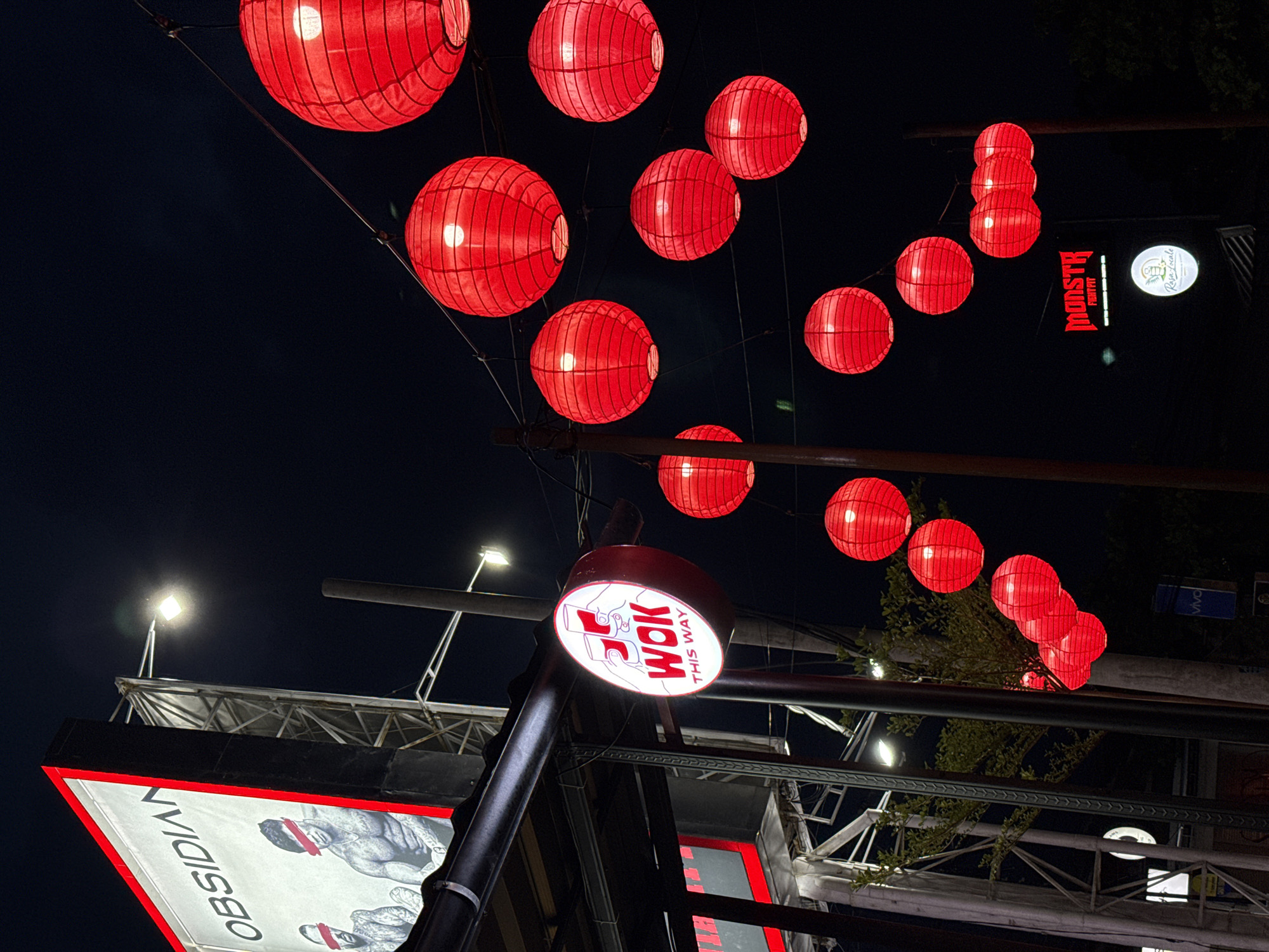 The image, taken at night in Munggu, Indonesia, shows a street scene decorated with red lanterns. These lanterns are strung above the street, creating a festive atmosphere. Signage for businesses such as "Monster Fight Fit", "Risa Locale", "Obsidian", and "Wok This Way" are visible, indicating a commercial area. The scene is illuminated by the lanterns and nearby streetlights. No people are visible in the frame. The environment consists of architectural structures and foliage.