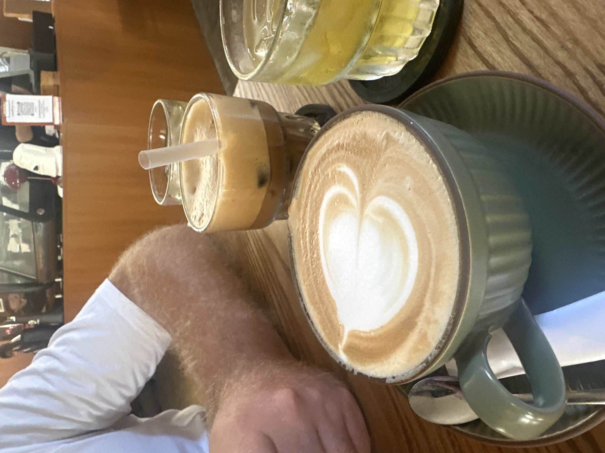 A first-person, angled close-up shows a table in what appears to be a cafe or restaurant. On the table are three beverages: a cappuccino with a heart design in the foam in a green cup with a matching saucer, an iced coffee with a straw in a tall glass, and a drink with a yellow liquid, possibly juice or lemonade, in a glass. Part of a person's arm and hand are visible, wearing a white sleeve. In the background, a shelf with items and what appears to be a storefront window are slightly blurred. The location is Bac Nha Trang Ward, Vietnam.