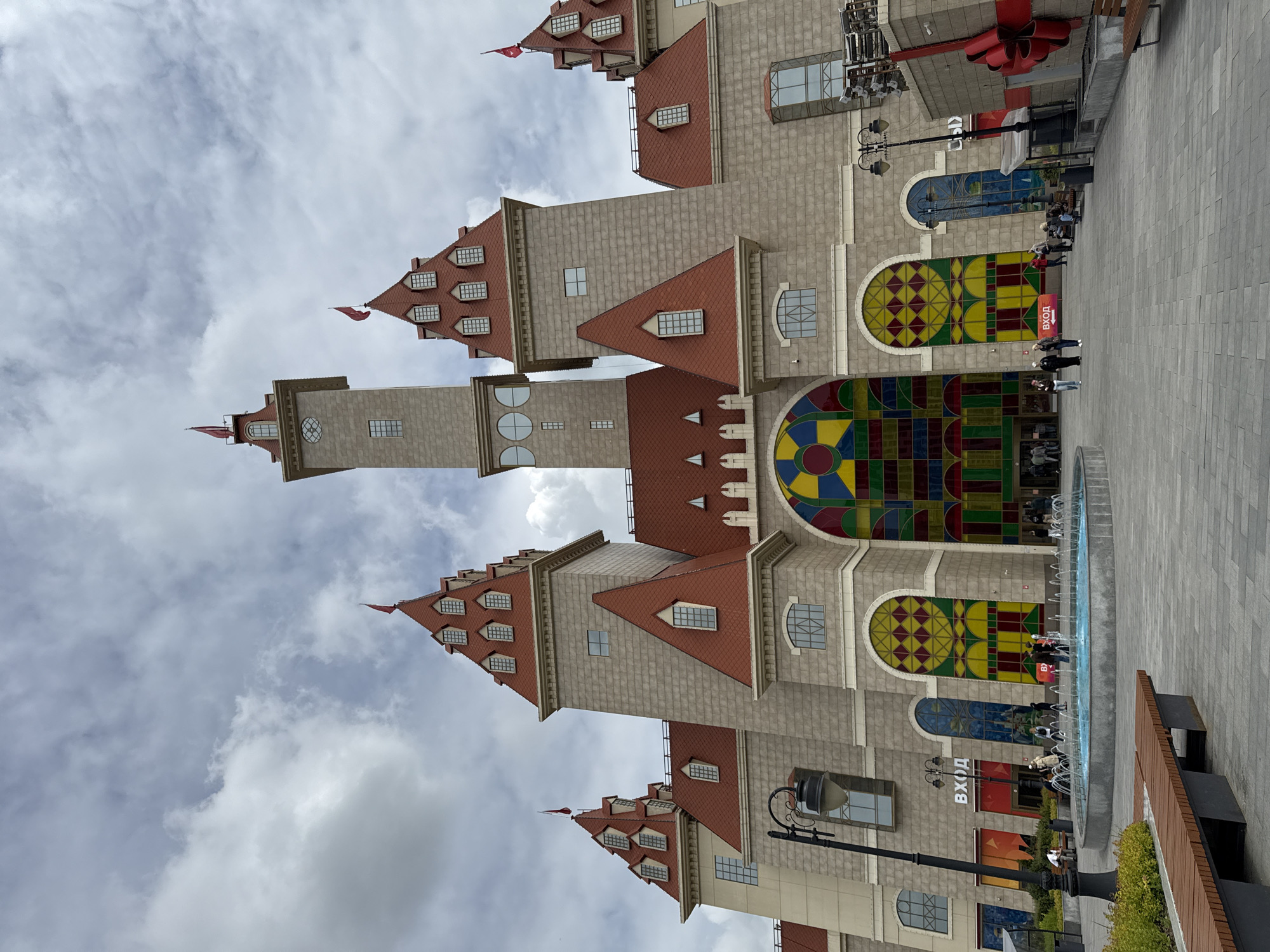 The image captures a large, ornate building resembling a modern castle or a themed entertainment complex in Moscow, Russia, under a cloudy sky. The structure features a grey stone-like facade, numerous towers and turrets, and prominent red-tiled pitched roofs with dormer windows. Some spires are topped with small red flags.
A key architectural highlight is the series of large, arched windows on the lower levels, adorned with vibrant, geometric stained-glass patterns in a kaleidoscope of colors, including reds, yellows, greens, and blues. One prominent window features a large circular pattern. The building also has various standard rectangular windows and round windows in one of its towers.
In front of the building, a wide paved pedestrian area extends, featuring a long, narrow fountain with multiple water jets. Several people are scattered across this area; some walk along the paving, while others gather near the fountain or entrances to the building. They appear to be casually dressed.
Other visible objects include dark-colored benches, a tall black lamppost with an elaborate design, and a large red decorative bow adorning the upper right section of the building.
The Russian word "ВХОД" (VKHOD), meaning "Entrance," is clearly visible on a red sign and above one of the building's entryways, confirming the building's function. The scene depicts a daytime setting with diffused light from the overcast conditions.