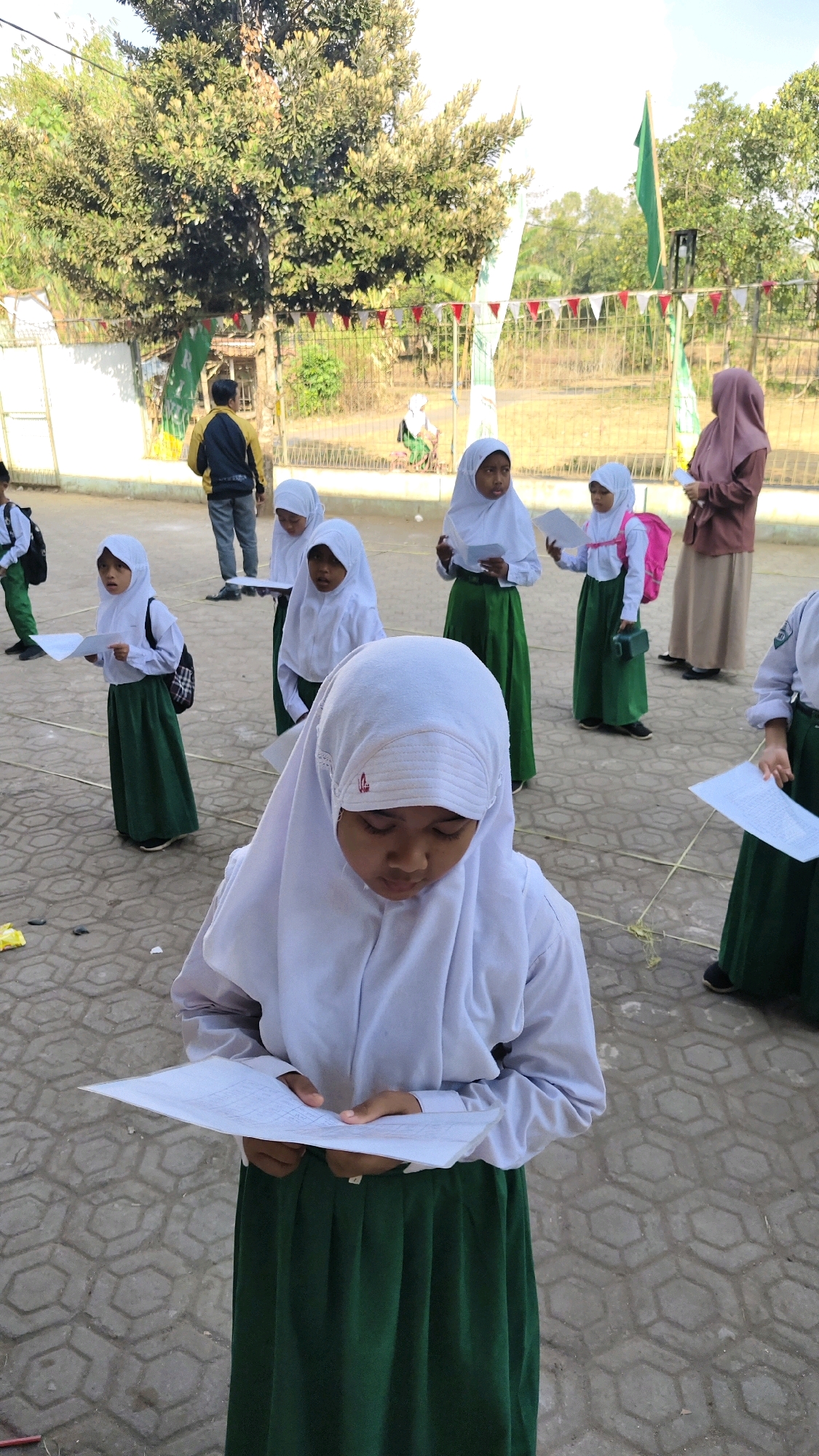 In Banyubunih, Indonesia, several young schoolgirls dressed in white hijabs and green skirts stand on a paved courtyard, each holding a piece of paper. A woman in a mauve hijab stands to the right, also holding paper. In the background, a man in a yellow and black jacket stands near a fence adorned with red and white pennant flags. Trees with lush green foliage are visible behind the fence, and a green flag flies from a pole. The overall scene suggests an outdoor school event or gathering.