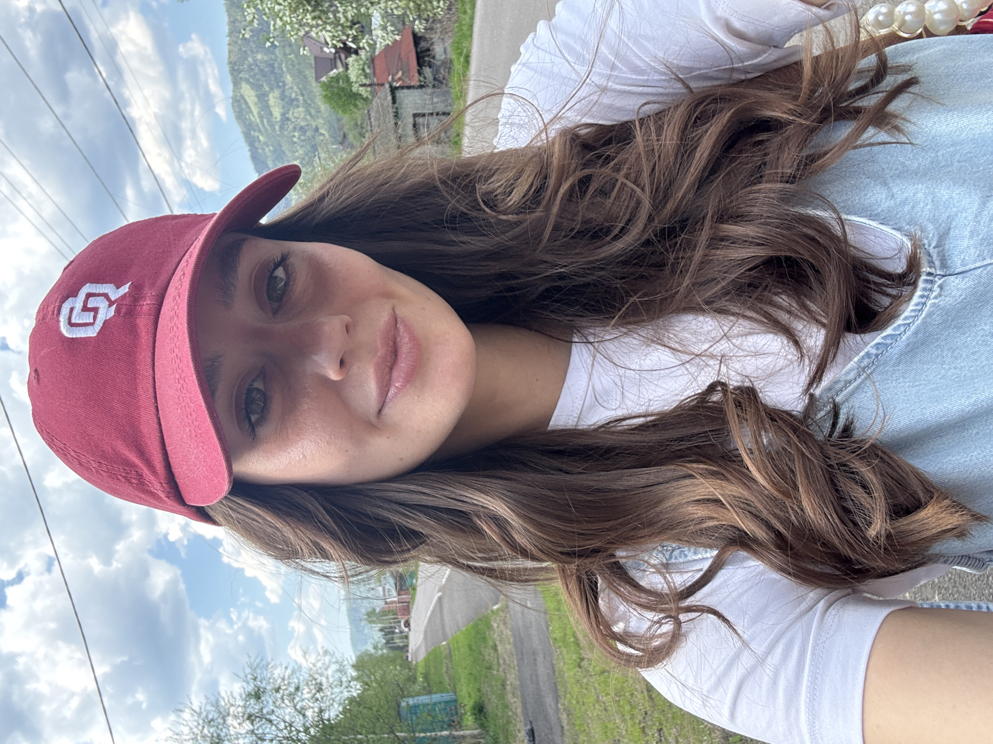 A woman with long, brown hair is posing for a selfie outdoors. She is wearing a burgundy baseball cap with a white logo, a white t-shirt, and a light-wash denim top. The background includes a paved road, grass, trees, and a distant mountain range under a cloudy sky. Based on the background's topography and structures, the location could potentially be Овсянка, Russia.