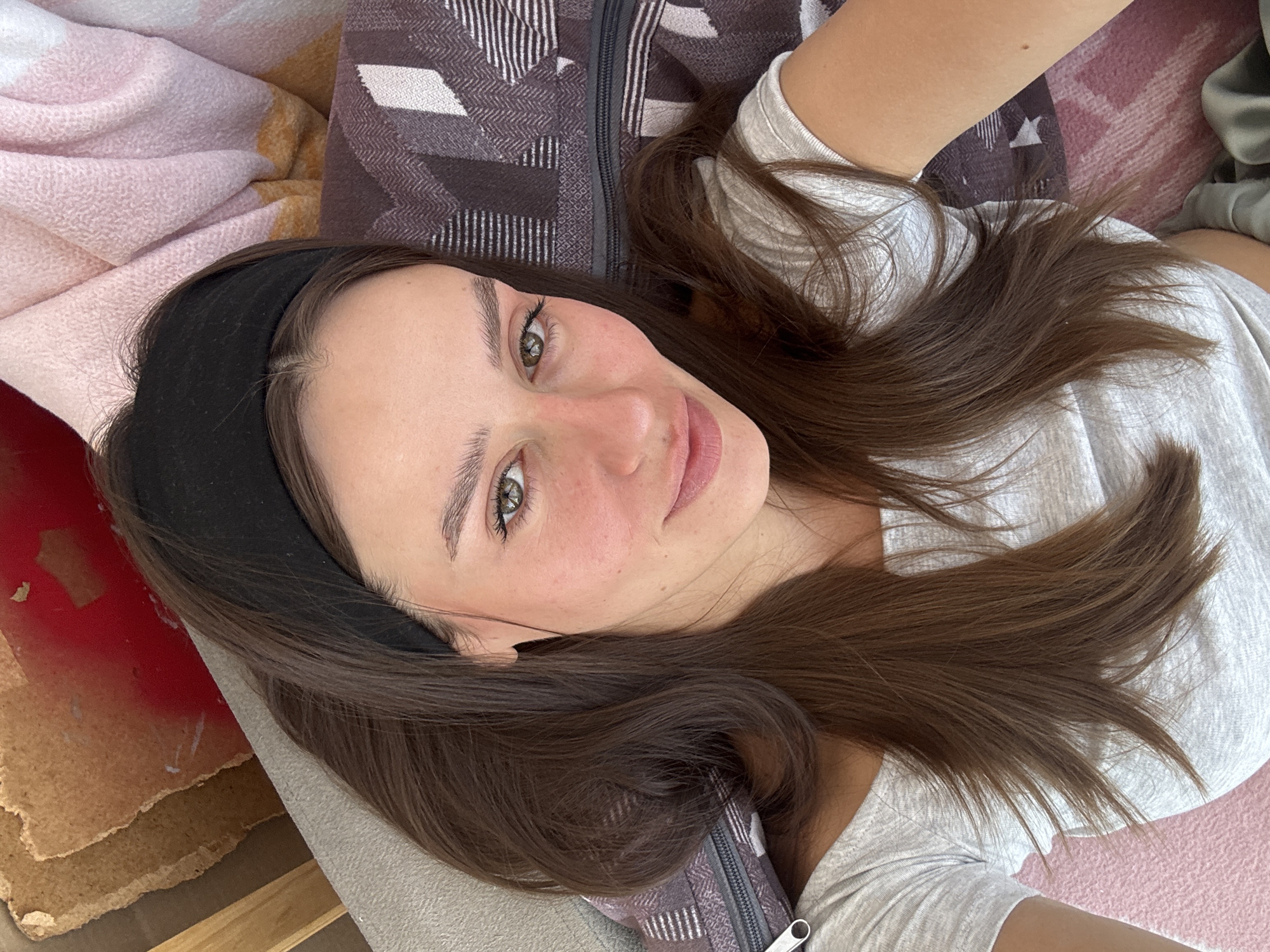 A young adult female with long, wavy dark brown hair, parted centrally and held back with a black headband, is depicted in a close-up selfie-style image. She has fair skin with a natural blush or redness across her cheeks and nose, and a small mole above her upper lip. Her eyes are a striking greenish-hazel, and her lips are slightly parted in a gentle smile, coated with a light pink or mauve lip color. She is wearing a light grey off-the-shoulder t-shirt.
The woman is positioned lying down, with her head resting on a light grey textured surface. To her immediate right, a dark purple or brown textured fabric with a distinct white/light geometric pattern (chevrons and diamond-like shapes) and a visible zipper is present, possibly a cushion cover or a patterned bag. Further to the right and above, a soft, light pink textured blanket with hints of white and orange-yellow is visible. In the upper left background, behind her head, there are fragmented layers of light brown and beige material, exposing a patch of red underneath, suggesting a worn or raw surface. A light pink textured surface is also visible in the lower left portion of the image. The scene is situated in Krasnoyarsk, Russia.