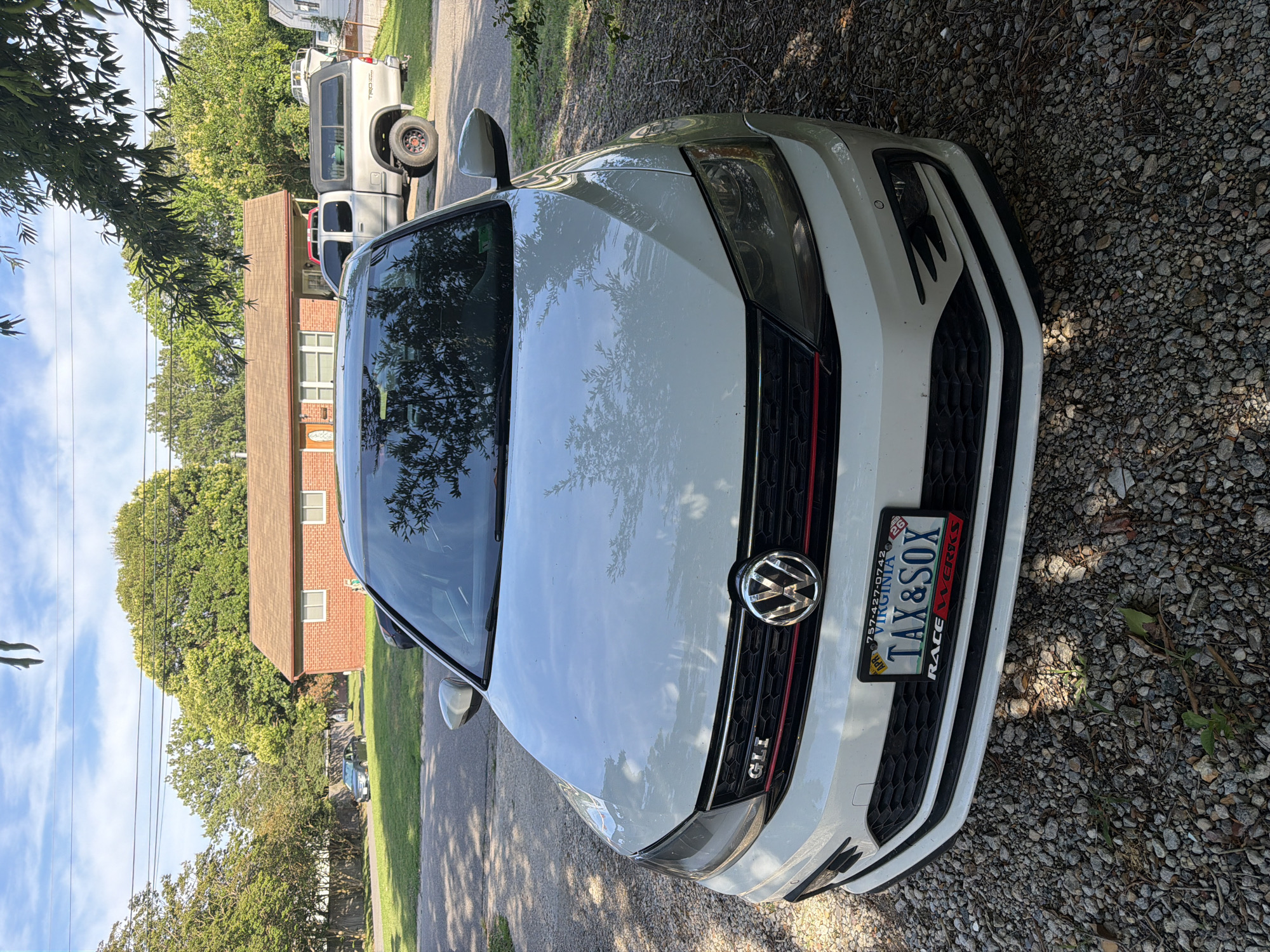 A white Volkswagen Jetta GLI is parked on a gravel driveway in front of a single-story brick house. The car's front is angled towards the camera, showing its license plate which reads "VIRGINIA TAX & SOX". The house has a brown roof and several windows. A silver pickup truck is parked to the right of the house, and a darker colored sedan is visible in the background on the left. The scene is illuminated by sunlight, casting shadows from trees onto the driveway and car. The location appears to be a residential neighborhood in Virginia Beach, USA.