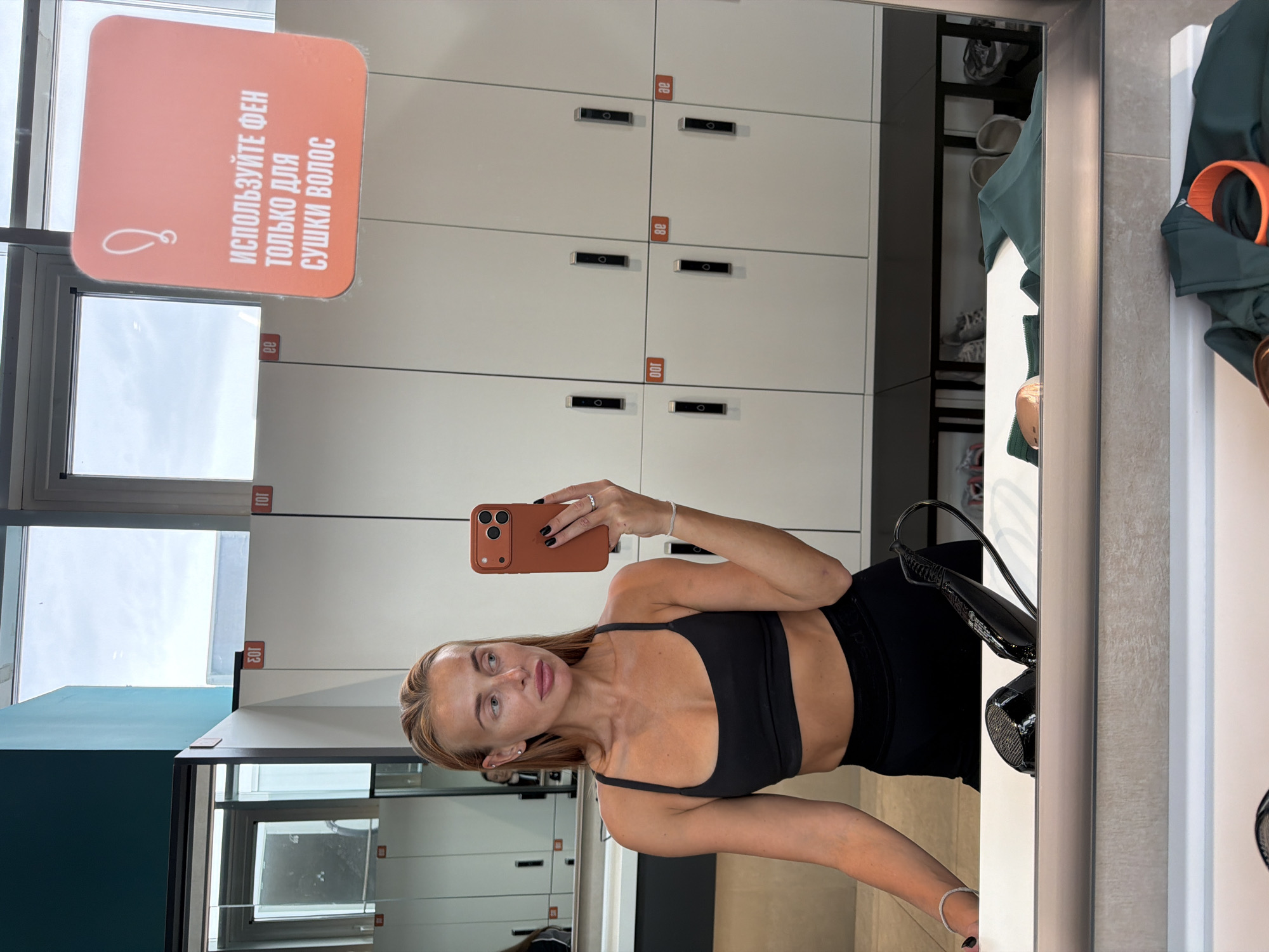 A young woman with blonde hair, wearing a black sports bra and black leggings, is taking a mirror selfie in a locker room. She is holding a smartphone in her right hand, which is covered by an orange case. The background consists of white lockers with digital locks. Above the lockers, there are windows and a sign in Russian that reads "USE THE HAIR DRYER ONLY FOR DRYING HAIR". There is a hairdryer visible in the mirror in the foreground. The locker room appears to be in a fitness facility in Krasnoyarsk, Russia, given the Cyrillic text on the sign.