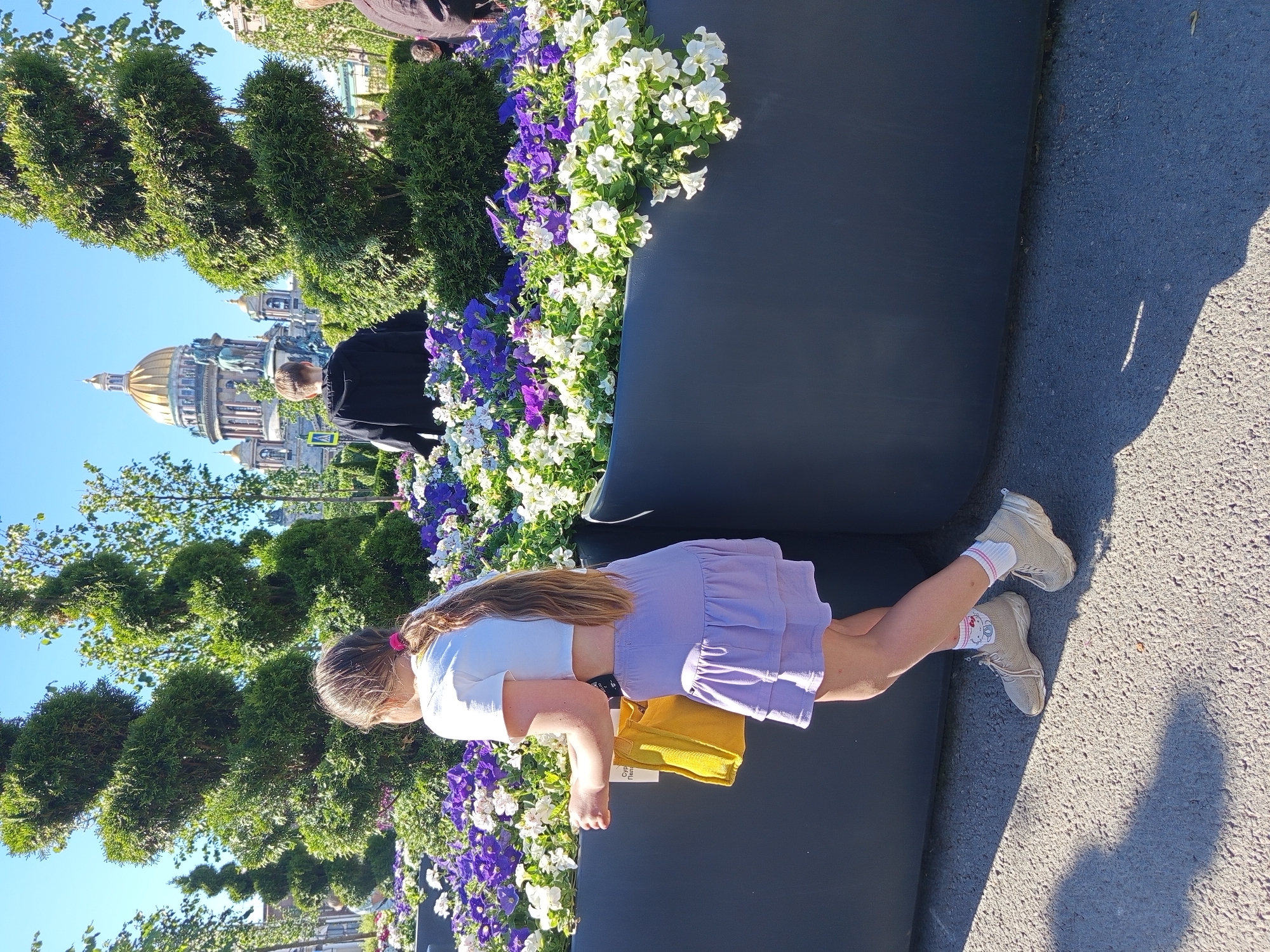 A girl in a white crop top and a lavender ruffled skirt is standing on asphalt. She has long, light brown hair pulled back into a ponytail with a pink elastic. She is holding a yellow bag in her left hand. She is wearing white socks with a Hello Kitty pattern and pink stripes, and beige sneakers. Behind her, a large dark planter box is filled with an abundant display of purple and white petunias. In the background, a man in a black hoodie faces away from the camera, and beyond him, the golden dome of St. Isaac's Cathedral rises against a clear blue sky. Spiral-shaped topiaries are visible on either side of the cathedral, framing the view. The scene appears to be outdoors on a sunny day.