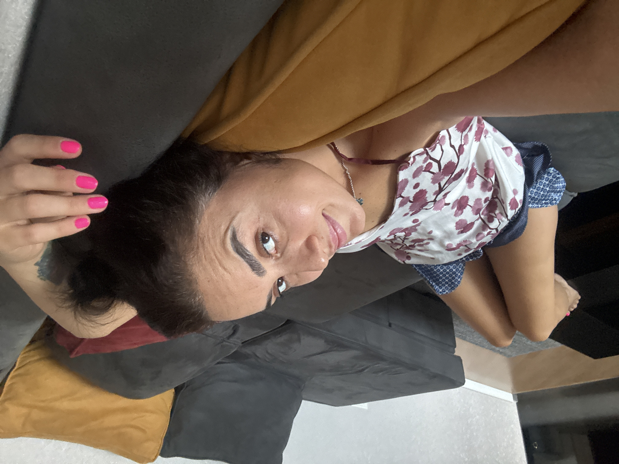 A woman is lying on a sectional sofa in a living room. She is facing the camera and has her right arm resting on a yellow pillow. Her legs are bent and her feet are visible in the foreground, with bright pink nail polish on her toes. She is wearing a white tank top with a floral pattern of pink flowers and green leaves. The sofa is dark grey, and there is a red pillow visible behind her. The background shows a wall and a glimpse of a floor. The environment appears to be indoors. The woman's face is partially visible, and she has dark hair. The scene suggests a casual and relaxed setting. The image appears to have been taken in Tbilisi, Georgia.