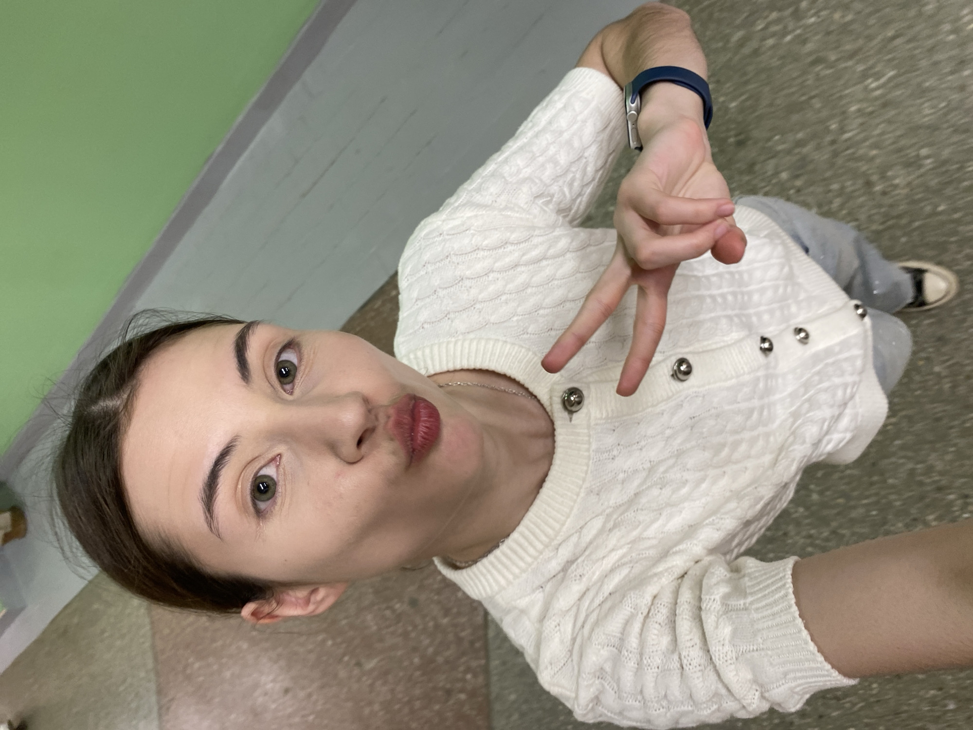 A young woman is taking a selfie indoors. She is wearing a white, textured cardigan with silver buttons and light-colored pants. She is making a kissing face and holding up two fingers in a peace sign. She has a blue smart watch on her left wrist. The background shows a wall with green paint and a floor with grey textured flooring. The location is likely a building interior, but not specifically identifiable as being within Votkinsk, Russia from the image alone.