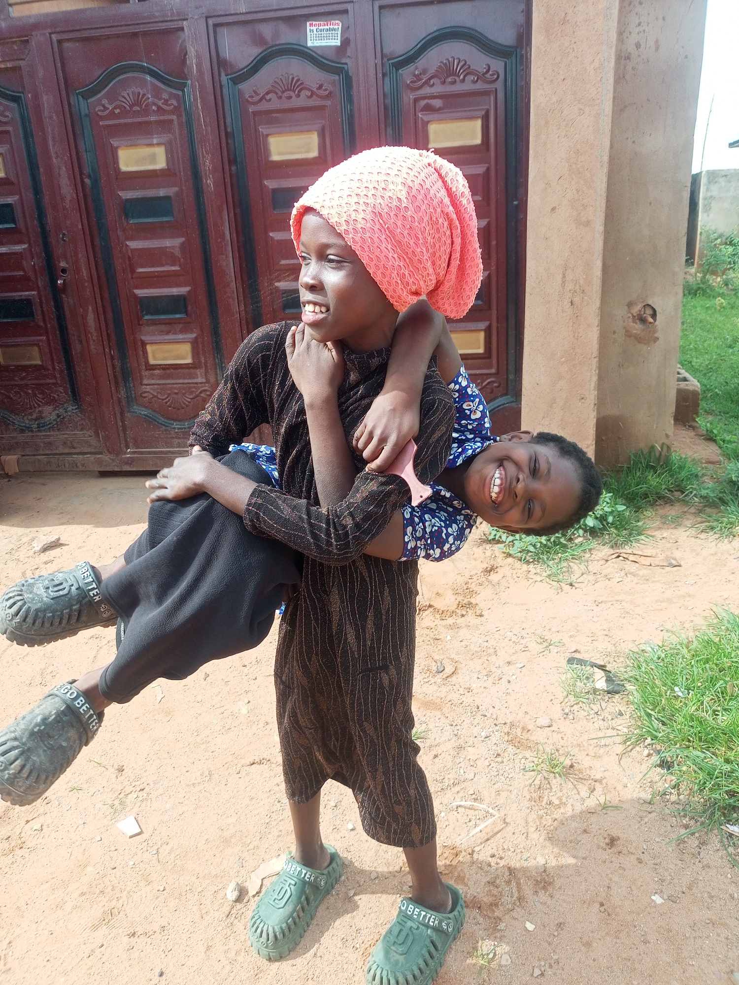 In the city of Tempure, Nigeria, two young girls are captured in a moment of playful interaction. One girl, wearing a pink headscarf and a long-sleeved patterned dress, lifts the other girl, who is smiling broadly and looking towards the camera. The second girl is dressed in a blue and white floral shirt. They are standing in a dirt area with some sparse green grass and a large, brown, double-doored gate in the background. The gate has decorative elements and a small red and white sign attached to it. One girl is wearing green clogs with the word "BETTER" visible on the strap, and the other is wearing similar clogs on her raised leg. The overall scene suggests an outdoor, casual setting.