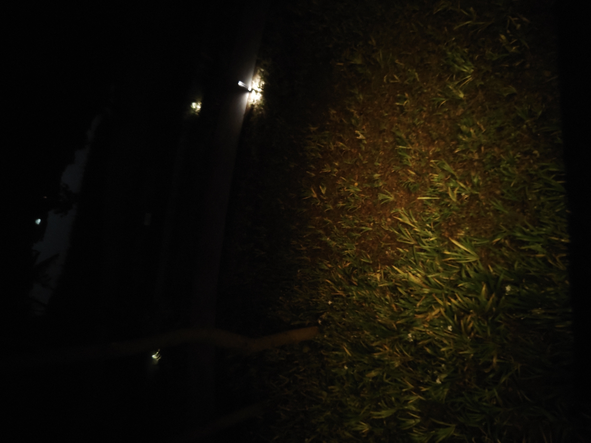 The image shows a dimly lit scene at night. On the right side of the image, there is an area of grass or low-lying vegetation. To the left, there is a dark, undefined area, possibly a road or pavement, with faint light sources illuminating the edge. A blurred, elongated shape is present, which may be a curb or a piece of debris, or a pipe. The location is identified as Chongqing High-tech Zone, China. No people, specific objects or actions are discernable in this image.