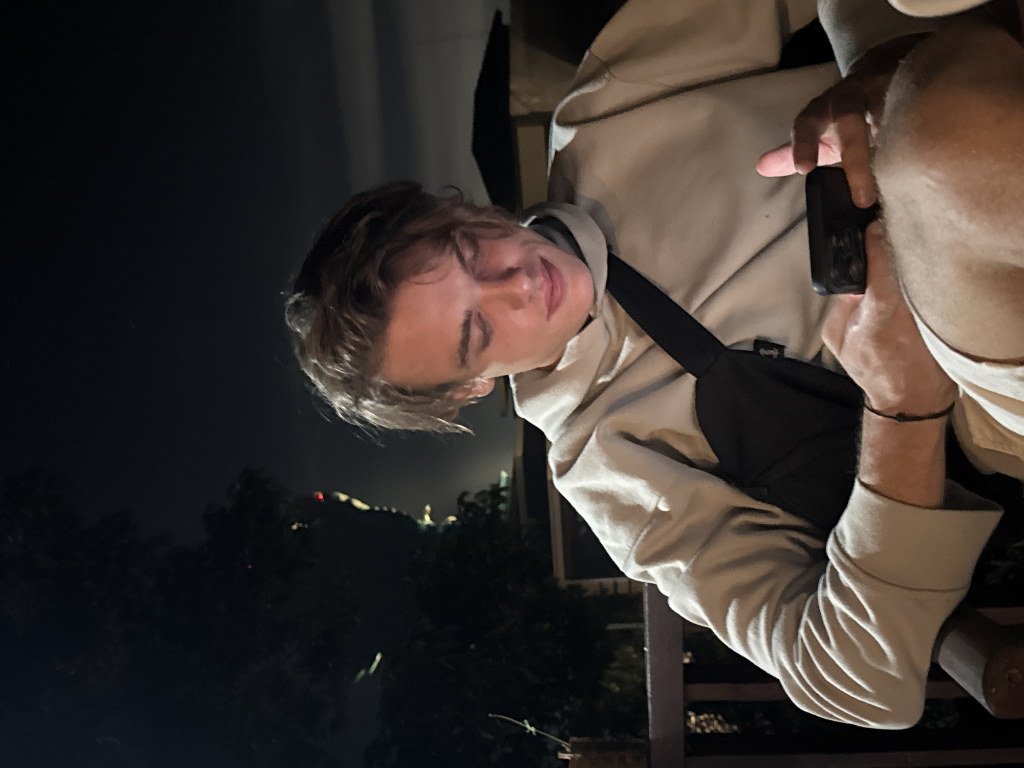 An outdoor, nighttime, medium shot captures a young individual looking at a smartphone from an elevated position in Kuta Selatan, Indonesia. The person, positioned in the right half of the frame, has light skin, light brown, wavy hair, and appears to be smiling or smirking, with eyes partially closed, suggesting amusement or focus on their device.
They are wearing a light beige or cream-colored hooded sweatshirt and a black sling bag across their chest. Their right hand is holding a dark-colored smartphone, with fingers slightly curled around it, indicating interaction. A thin, dark bracelet is visible on their left wrist. A wooden railing is partially visible in the lower right foreground.
The background is dark, characteristic of nighttime. Distant city lights or building lights are visible on a lower horizon, suggesting a view over a developed area from a height. Dark tree silhouettes are discernible between the foreground subject and the distant lights. The overall setting implies a balcony or terrace overlooking a town or city at night. No discernible text is visible in the image.