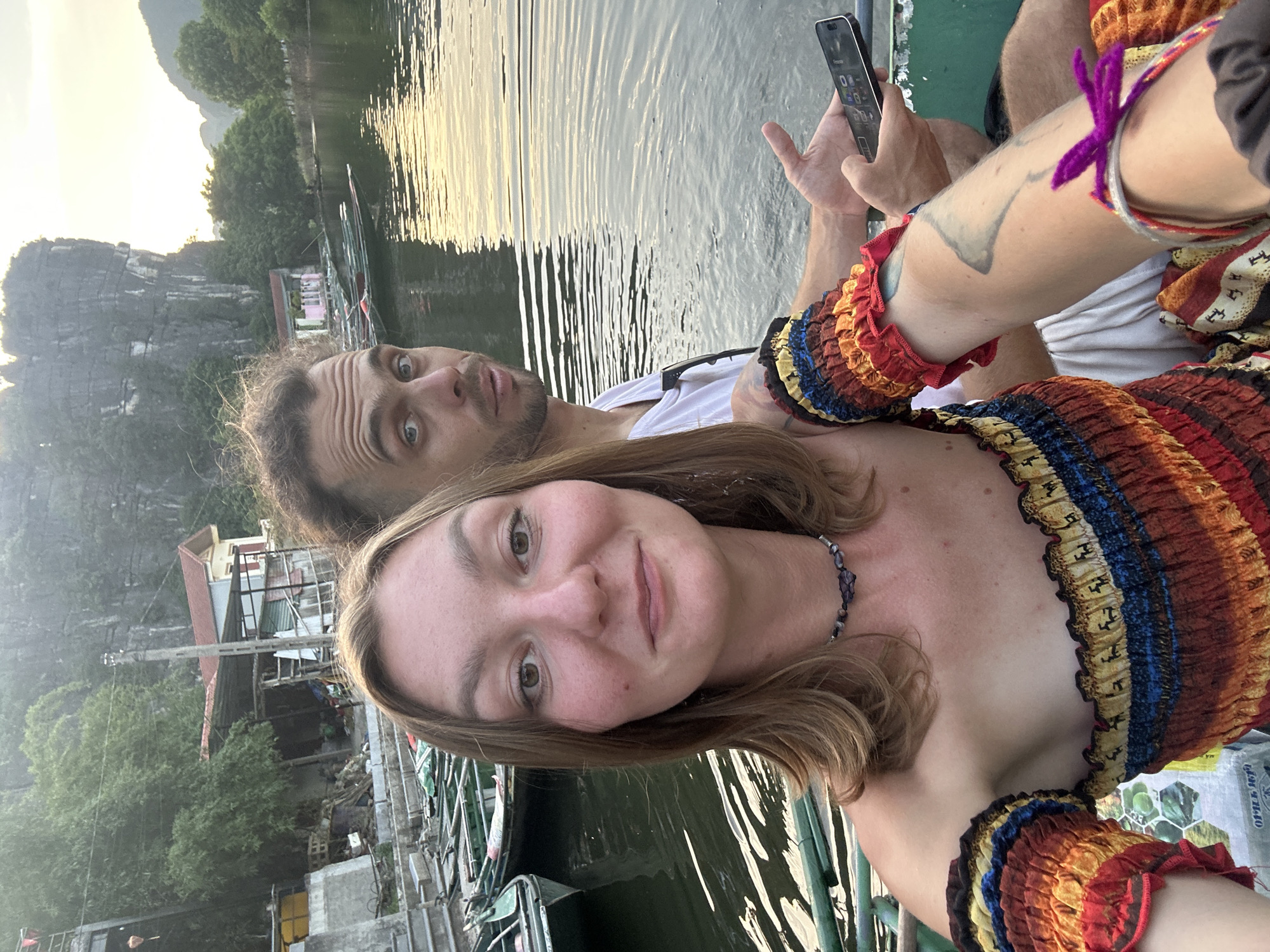 In Nam Hoa Lư Ward, Vietnam, two individuals, a woman and a man, are seated in a boat on a body of water. The woman is in the foreground, facing the camera, and is wearing a colorful off-the-shoulder top with ruffled sleeves and a beaded necklace. She has medium-length brown hair and is smiling. The man is behind her, looking towards the camera with a surprised expression. He has short brown hair and is wearing a white t-shirt. He is holding a smartphone in his right hand. The background shows a serene landscape with limestone karst mountains, lush green trees, and several other boats in the water. The sun appears to be setting, casting a warm glow on the scene. The water is calm and reflects the sky and surrounding environment.