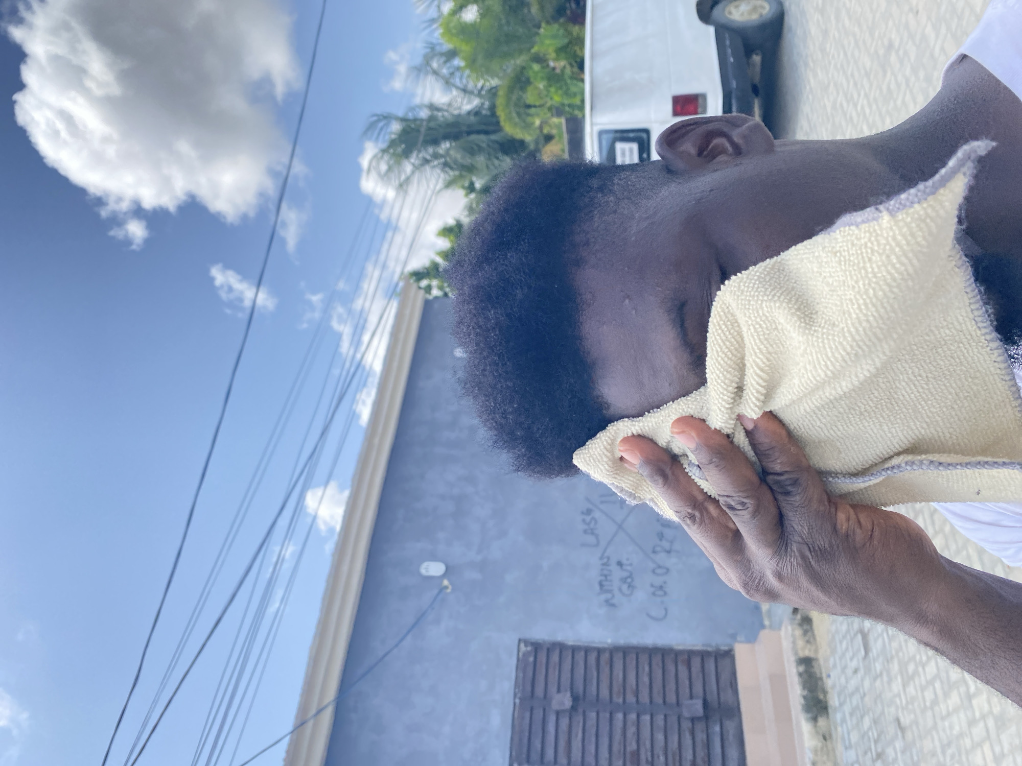 The image, rotated correctly, captures a close-up, side profile of a person with dark skin, whose face is mostly obscured by a light-colored towel held in their right hand. The person has short, dark, curly hair and is wearing a white garment.
The scene is outdoors on a bright, sunny day, indicated by the clear blue sky dotted with scattered white, fluffy clouds. Power lines crisscross the upper part of the frame. In the background, a light blue/grey building wall is visible, with a dark, shuttered window or door towards the bottom. Graffiti on this wall includes the words "WITHIN LASS" and "G.P.T.," an 'X' symbol, and the date "C.06.07.22."
To the far right, lush green foliage, possibly palm trees, and the rear section of a white vehicle (likely a van or bus) with a red taillight and a visible wheel are present. The ground appears to be paved with light-colored, patterned tiles or cobblestones. The person is engaged in the act of wiping their face, suggesting a response to heat, sweat, or personal hygiene in an outdoor setting.