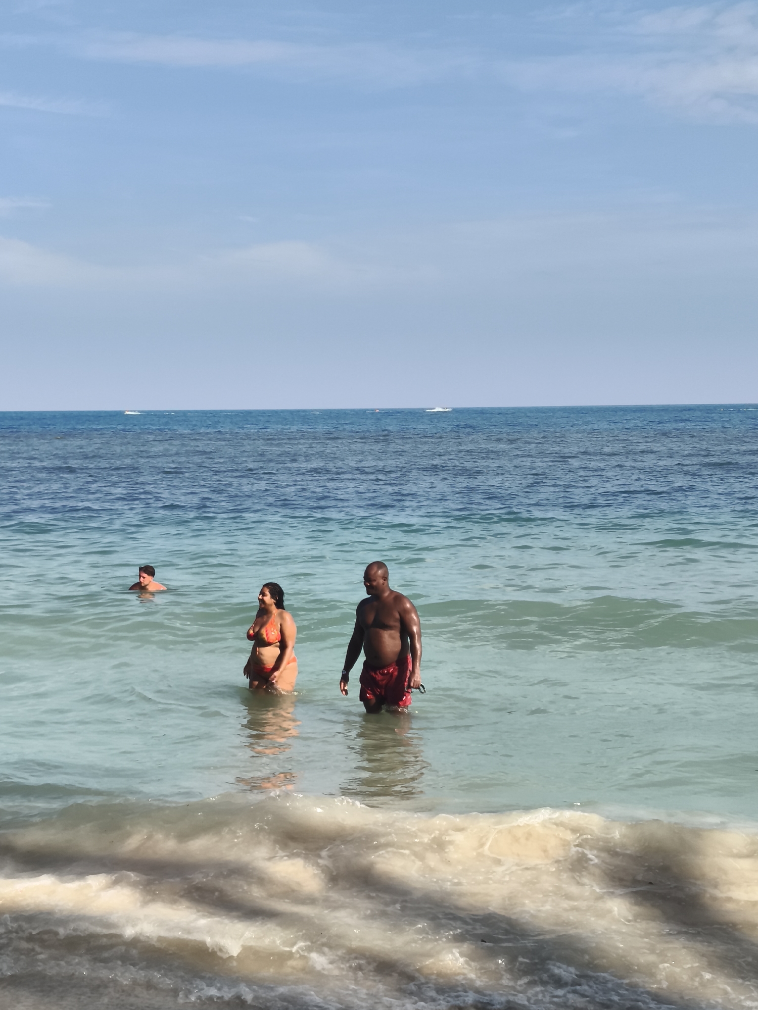 The image depicts a tropical beach scene with three individuals in the water. In the foreground, a man with dark skin and red swim trunks stands facing forward. To his left, a woman in an orange bikini is also facing forward, with her body partially submerged in the water. Further back and to the left, a person wearing a snorkel mask is visible with their head above water. The water is a clear turquoise color, with gentle waves breaking on the shore in the immediate foreground. The ocean extends to the horizon, where it meets a clear blue sky. Two small boats are visible in the distance on the water. The overall impression is a sunny day at a beach, likely in a location with calm, clear waters. The context of Ko Samui District, Thailand, suggests a warm, tropical climate.