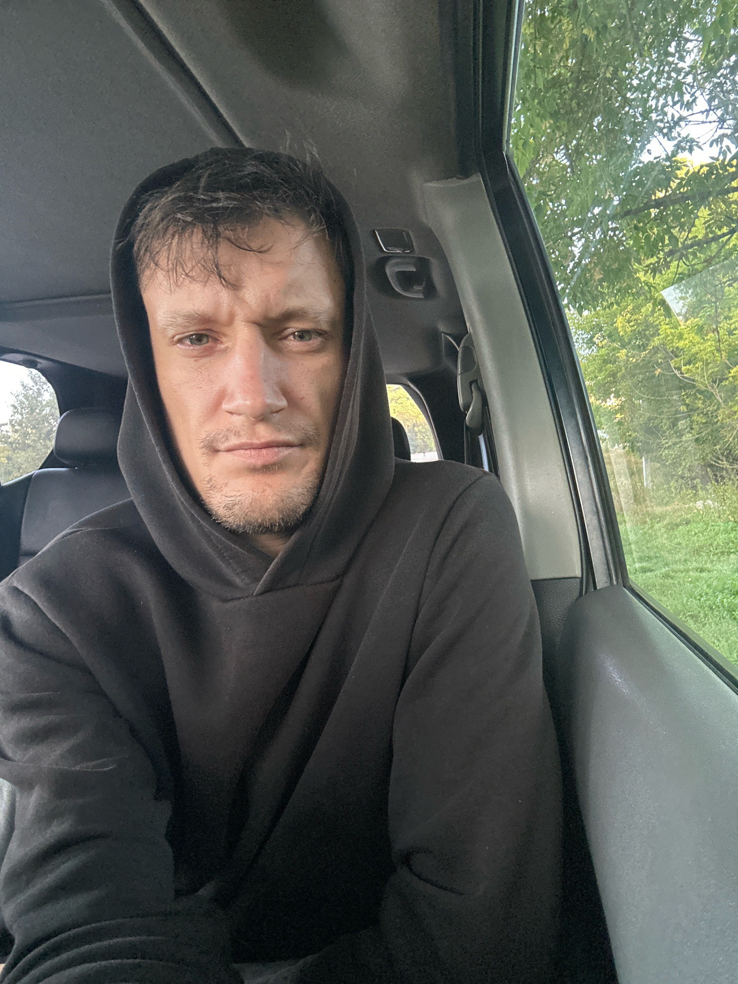 A selfie of a man in a black hoodie is taken from inside a vehicle. The man has short, disheveled brown hair and a light beard. His eyes are looking directly at the camera with a neutral expression. The interior of the vehicle appears to be gray and black, with a black leather seat visible behind him. The window on the right side of the vehicle shows a blurred background of green trees and grass, suggesting an outdoor, possibly rural or suburban, setting. The lighting suggests it is daytime, with natural light coming from the window and possibly a sunroof. There are no other people or significant objects visible in the frame. The location is identified as Makiv, Ukraine.