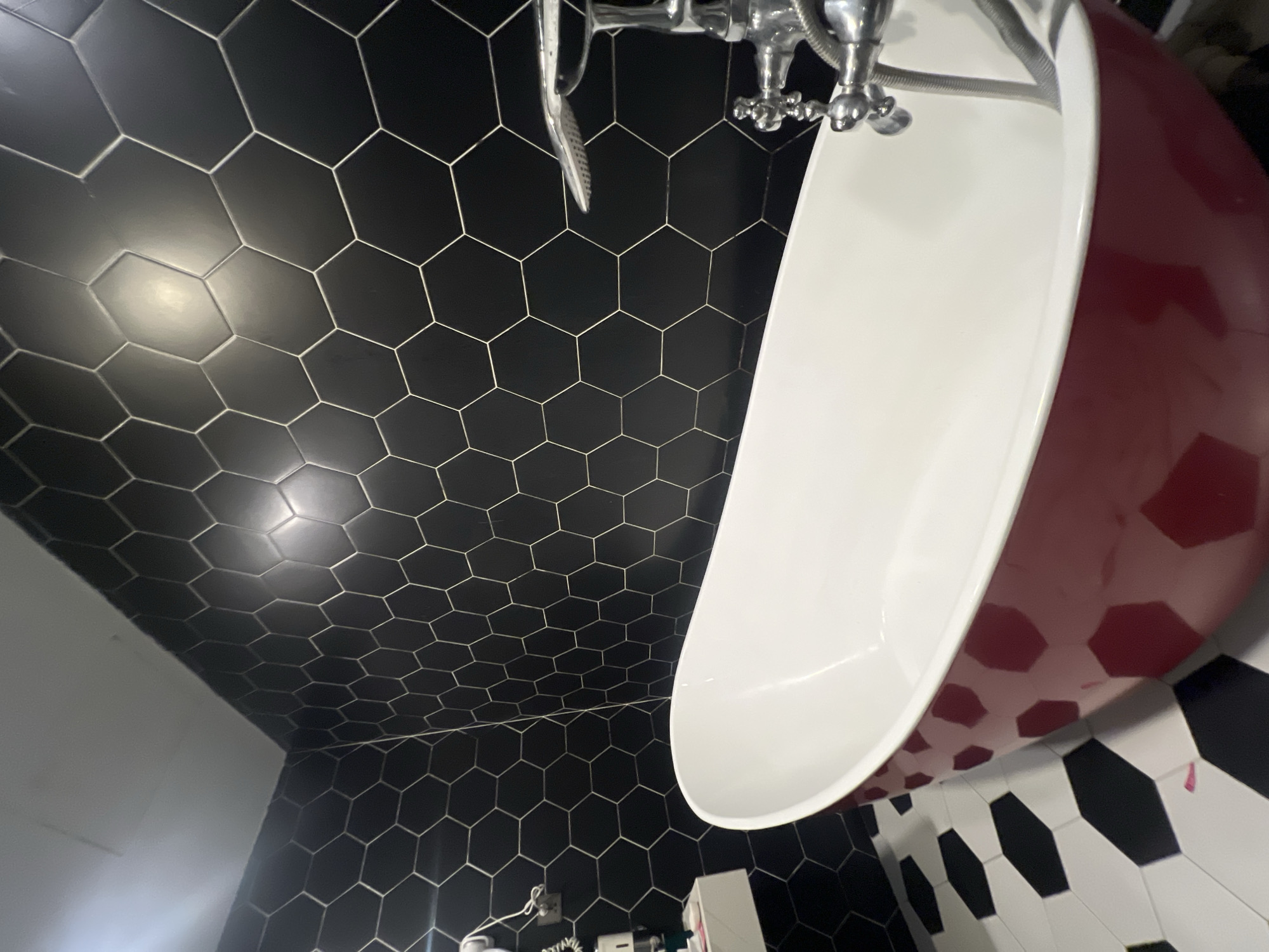 A bathroom features a red bathtub with a white interior, placed against a wall tiled with black hexagons and white grout. The floor is tiled with a pattern of alternating black and white elongated hexagons. A silver faucet and showerhead are visible near the rim of the bathtub. On the left side of the image, a white shelf is mounted on the wall, holding a hairdryer and other toiletries. The scene is captured indoors, likely in a residential setting. The image does not provide any specific information or visual cues to pinpoint its location to Hà Nội, Vietnam.