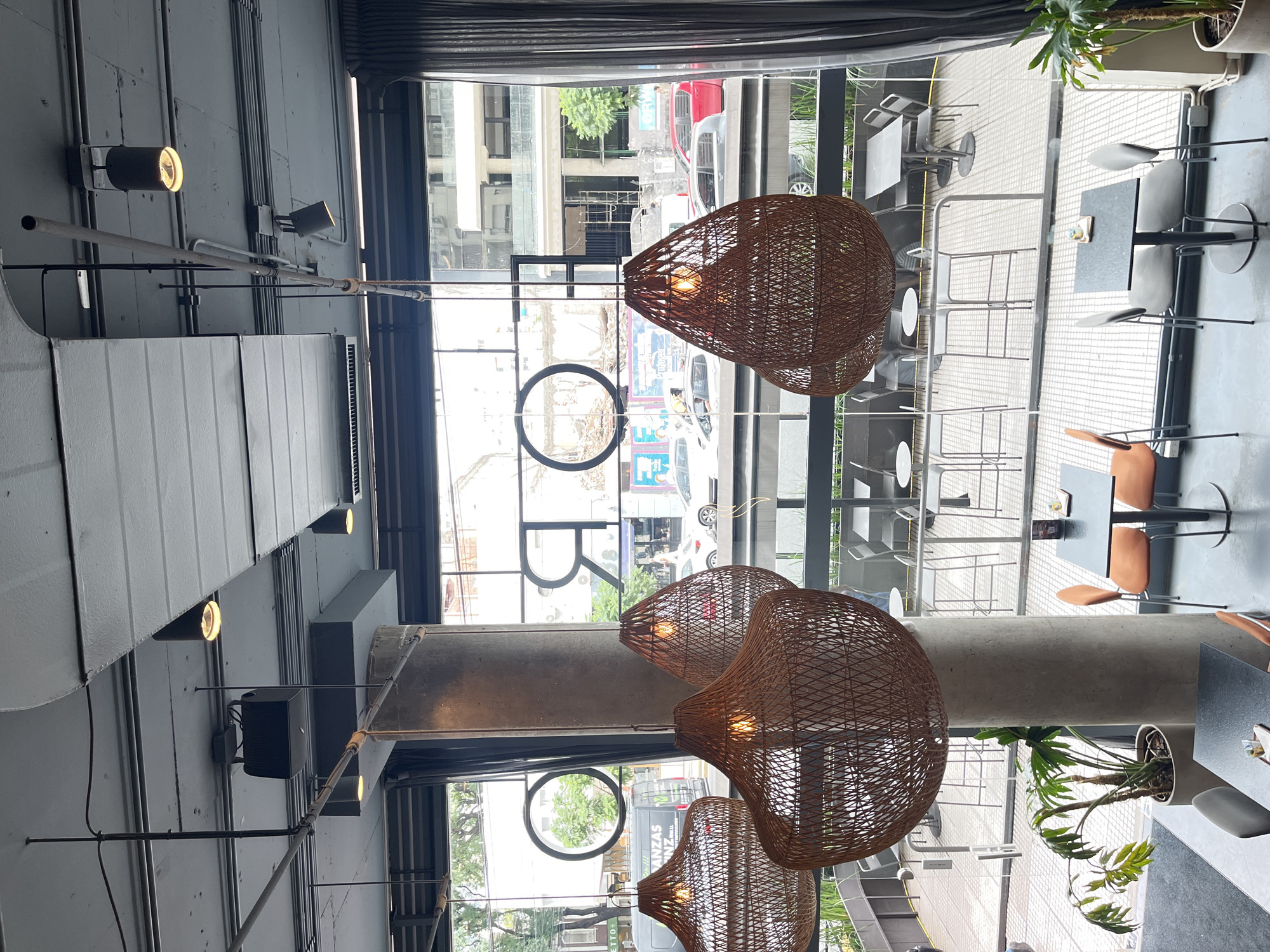 The image depicts the interior of a restaurant in Buenos Aires, Argentina. Several large, wicker pendant lamps hang from the ceiling. A tall window with large, black lettering "O B R" overlooks a street scene with cars and buildings. Outside, tables and chairs are visible on a patio. Inside, potted plants add a touch of greenery. The interior design features an industrial aesthetic with exposed pipes and concrete pillars. There are no people visible in the scene.