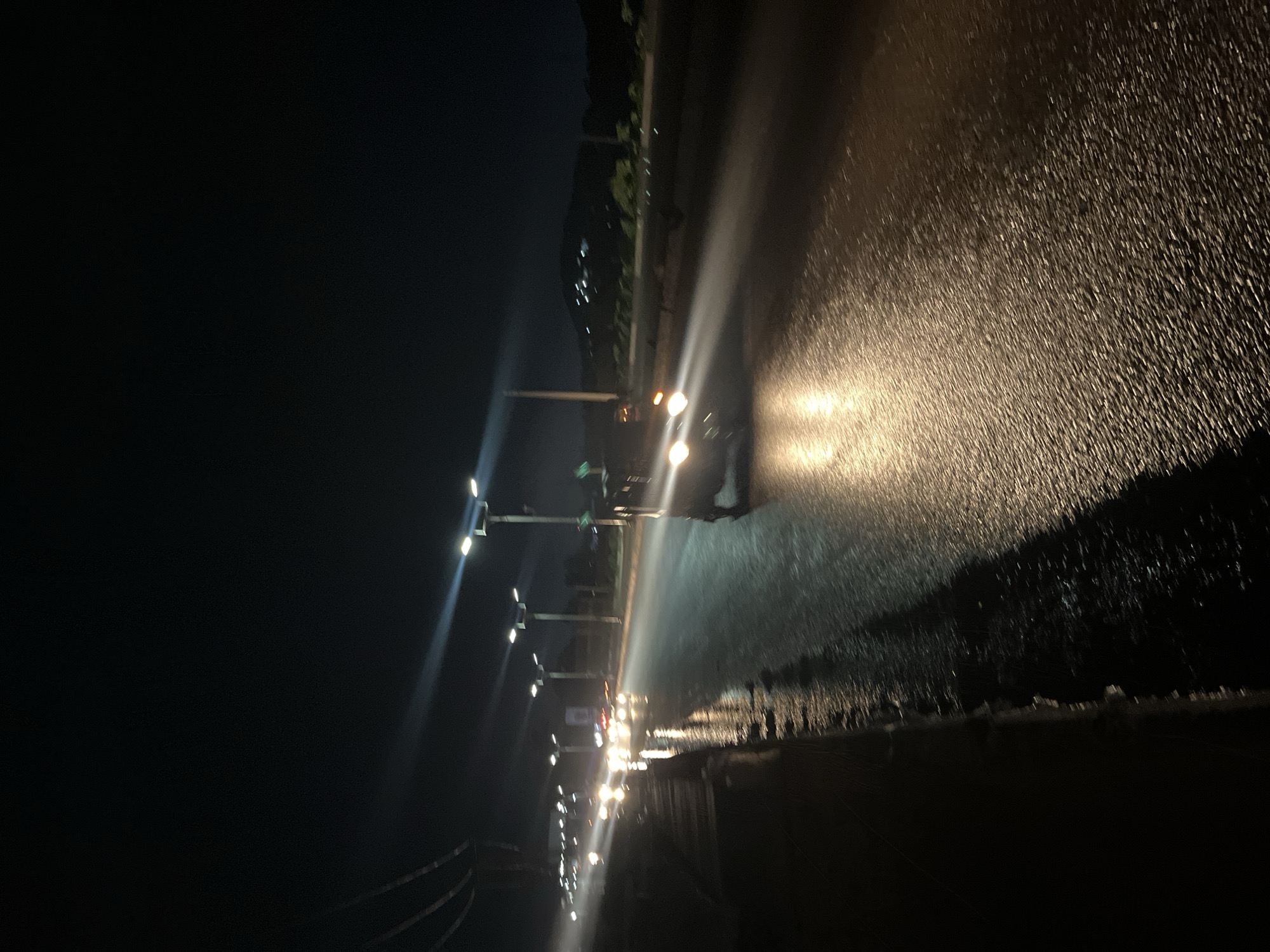 A night-time, low-light image captures a street scene in Gura Top, Nigeria. The road surface appears wet and reflective, likely due to recent rain. Streetlights line the right side of the road, casting bright beams onto the wet pavement, creating streaks of light. Several vehicles are visible on the road, their headlights illuminating the scene. One vehicle in the foreground, partially visible on the right, has its headlights on, creating a strong glare. Other vehicles are seen in the distance, with their lights appearing as bright blurs on the wet road. The background is dark, with a faint silhouette of hills or mountains visible in the distance, dotted with lights that suggest a settlement. The overall impression is of a busy road at night during or after rainfall.