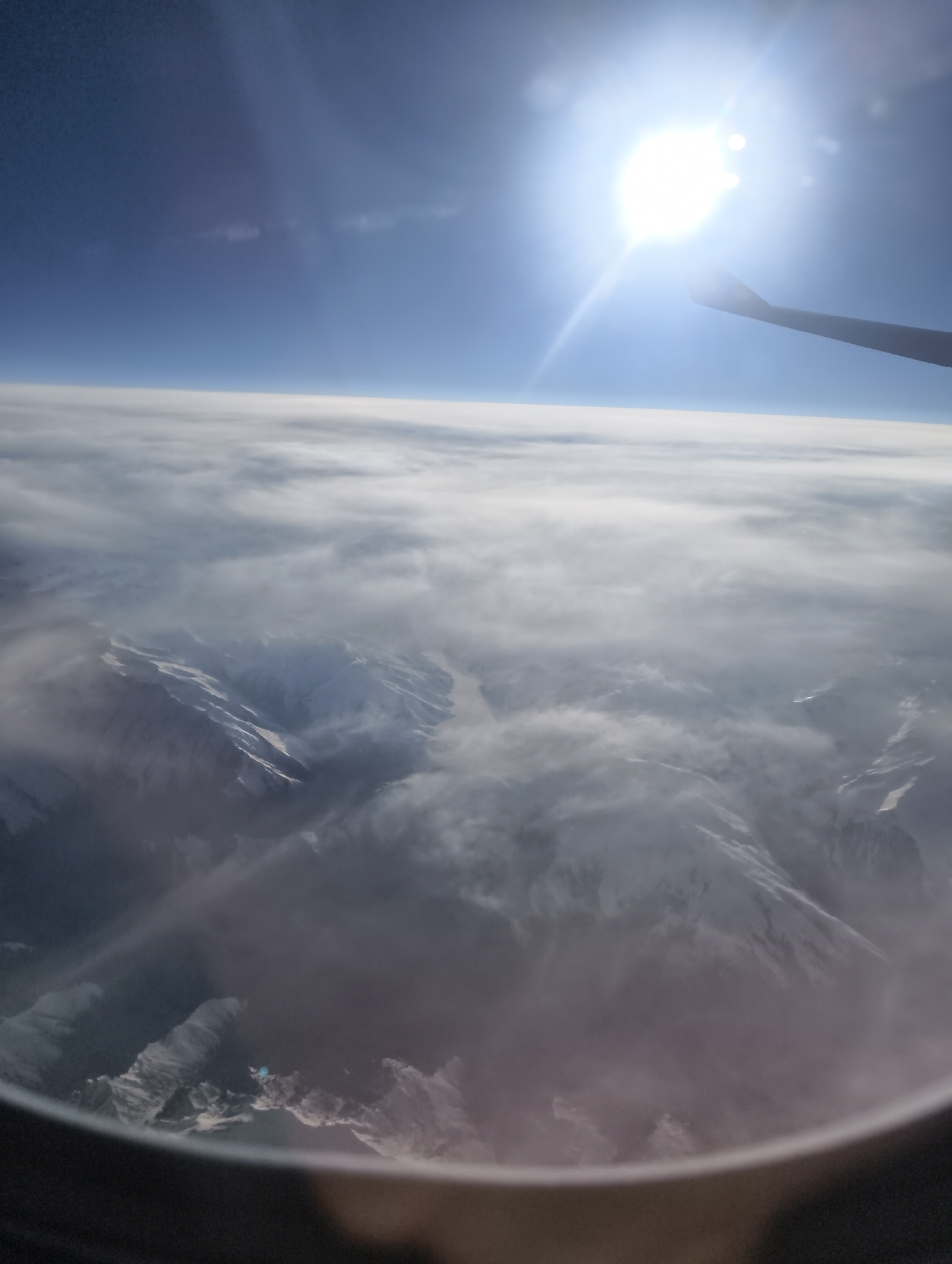A view from an airplane window shows a landscape of snow-covered mountains partially obscured by a thick layer of clouds. Above the clouds, a bright sun shines in a clear blue sky. Part of the airplane wing is visible in the upper right. The lower portion of the window frame is visible in the lower portion of the frame. The location and city are unknown.
