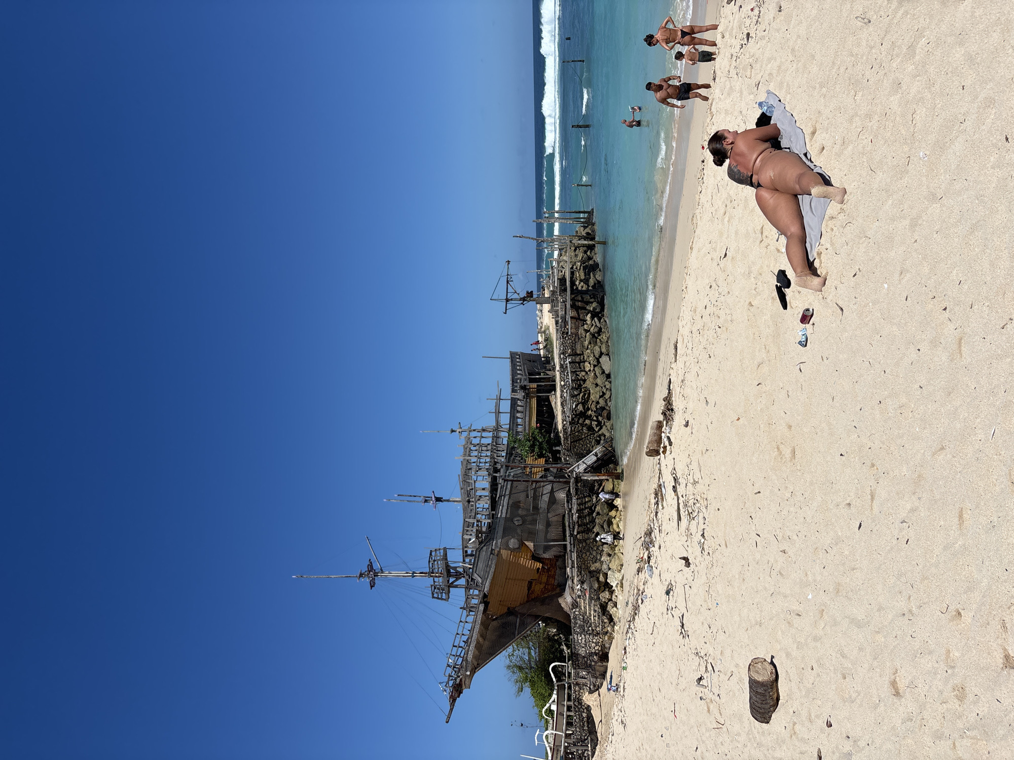 The scene depicts a beach in Kuta Selatan, Indonesia. A large wooden pirate ship structure sits on the beach, near a rocky pier. In the distance, people are swimming and standing in the ocean. On the right side of the image, a woman is lying on a towel on the sand. Debris is scattered on the beach around the ship and woman. The sky is clear and blue.