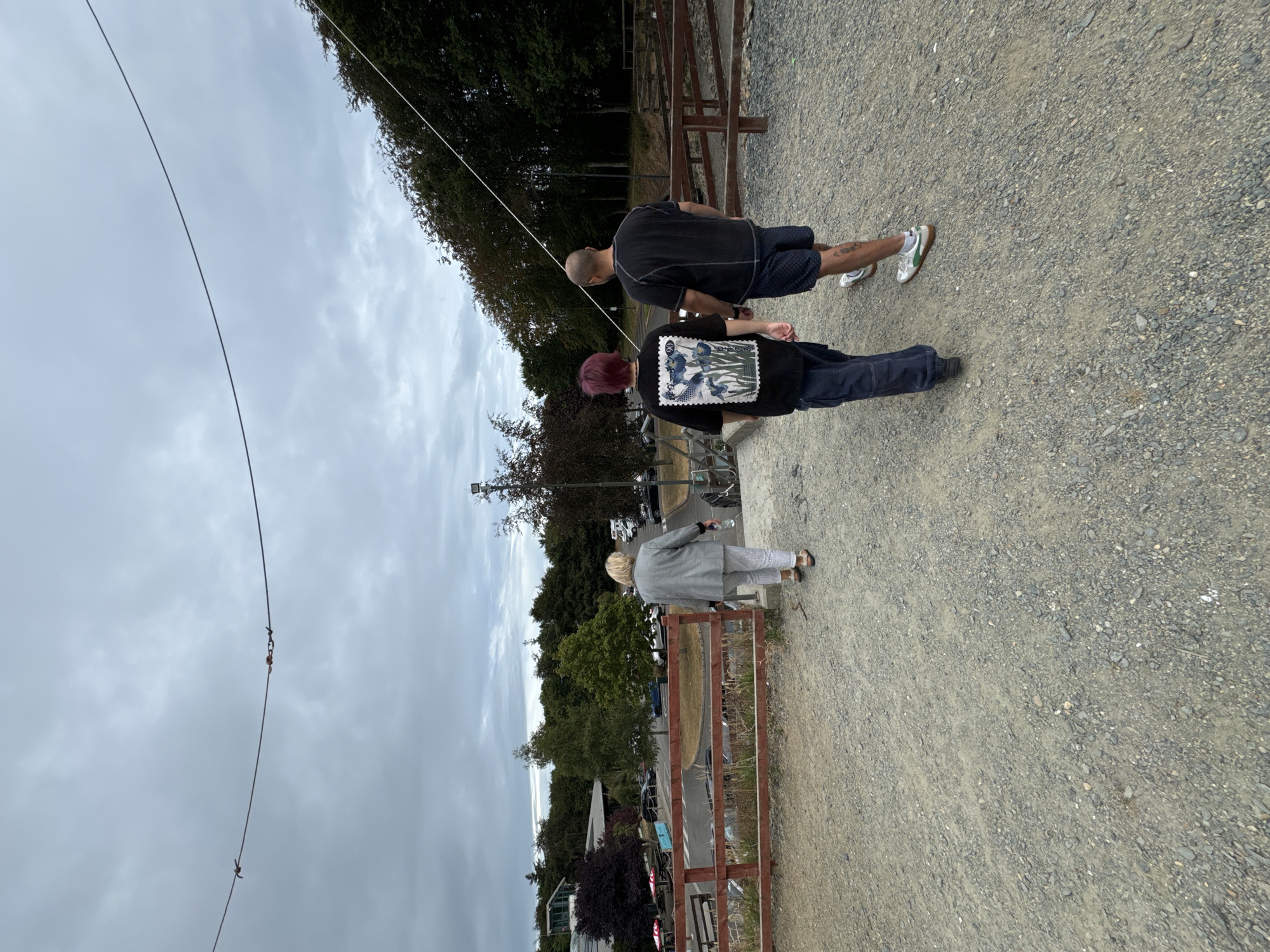Three individuals are walking away from the camera down a gravel path in Courtown, Ireland. The person on the left, a woman with blonde hair, is wearing a grey patterned jacket and striped pants. The person in the middle, with red hair, is wearing a black t-shirt with a floral design and dark pants. The person on the right, a man, is wearing a black t-shirt and patterned shorts. They are walking towards a wooden fence and a parking lot with cars visible in the background. Trees and buildings are also present in the surrounding environment. The sky is overcast.