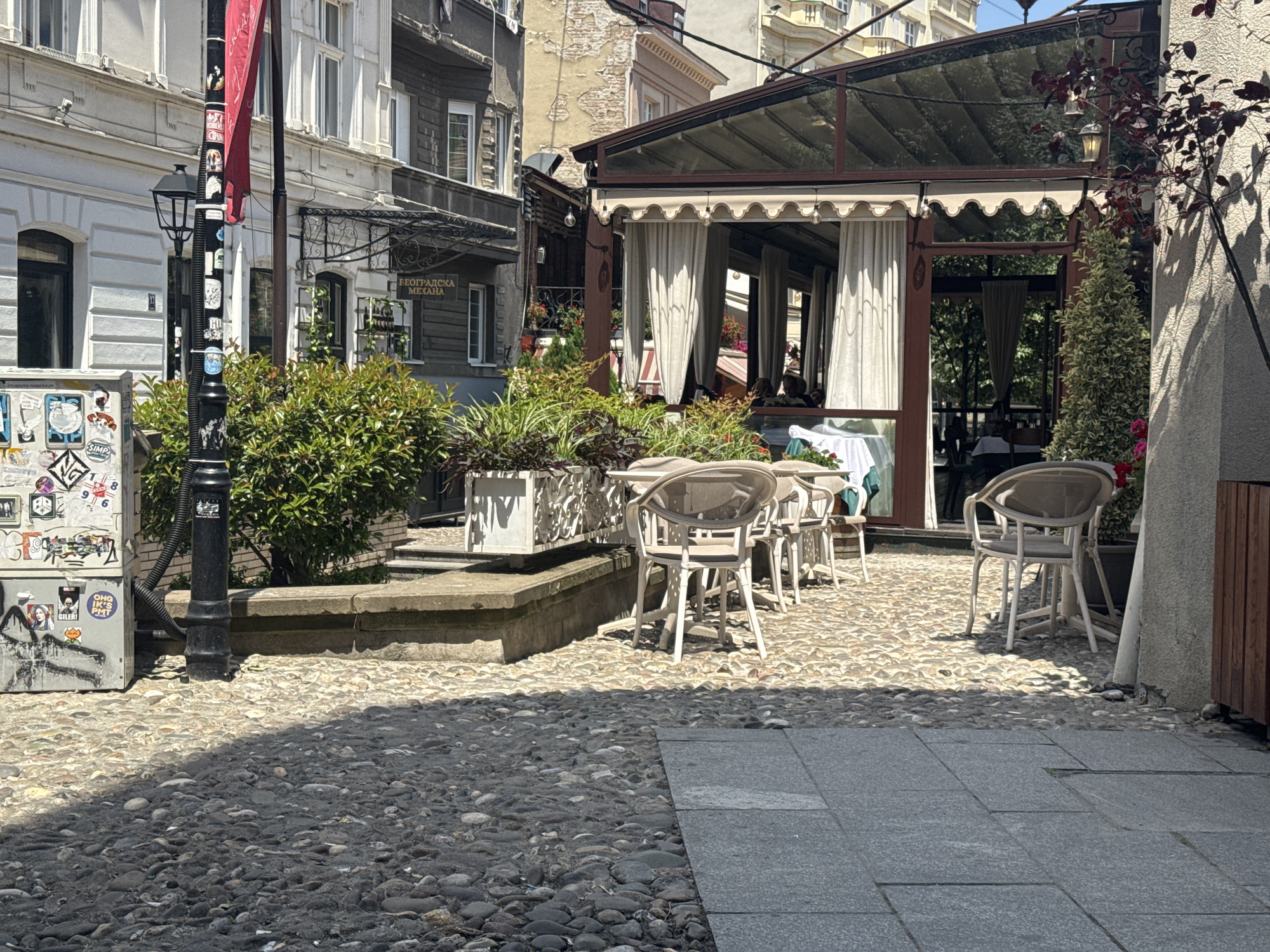 The image depicts a charming outdoor seating area of a restaurant, likely situated in the historic Skadarlija district of Belgrade, Serbia. The ground is paved with cobblestones, and to the right, a section of modern grey tiles is visible. A covered outdoor dining area with white curtains is present, with tables and chairs set up for patrons. Green foliage is strategically placed in planters, adding to the ambiance. A lamppost adorned with stickers stands prominently, and graffiti is visible on a utility box to its left. In the background, traditional Belgrade architecture is evident, with buildings showcasing distinct European styles. The scene appears to be captured on a sunny day, with shadows cast by the overhead structures and vegetation.
