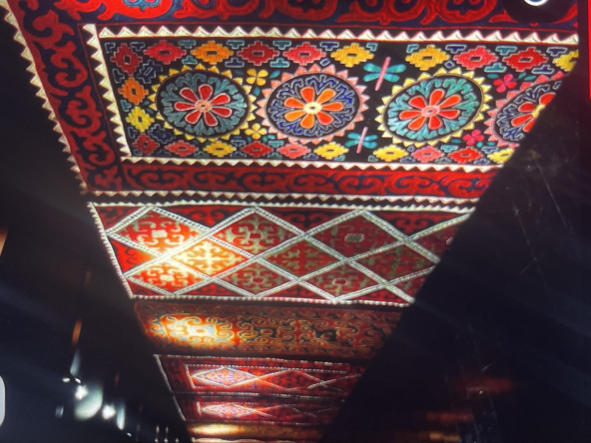 A wide-angle shot shows a collection of intricately patterned textiles, possibly suzanis or carpets, displayed vertically in a dimly lit exhibition space. The textiles are rich in color, featuring geometric designs and floral motifs in vibrant reds, blues, yellows, and greens against dark backgrounds. They appear to be illuminated by overhead lights, creating a dramatic effect. The perspective is from a low angle, looking up and to the right, emphasizing the scale and detail of the textiles. In the foreground, a dark, reflective surface, likely a floor, obscures much of the lower portion of the image. The overall impression is one of cultural display and artistic exhibition, possibly at a museum or gallery. This scene is consistent with displays found at the Kyrgyz State Museum of Fine Arts in Bishkek, Kyrgyzstan, which houses a significant collection of traditional Kyrgyz textiles.