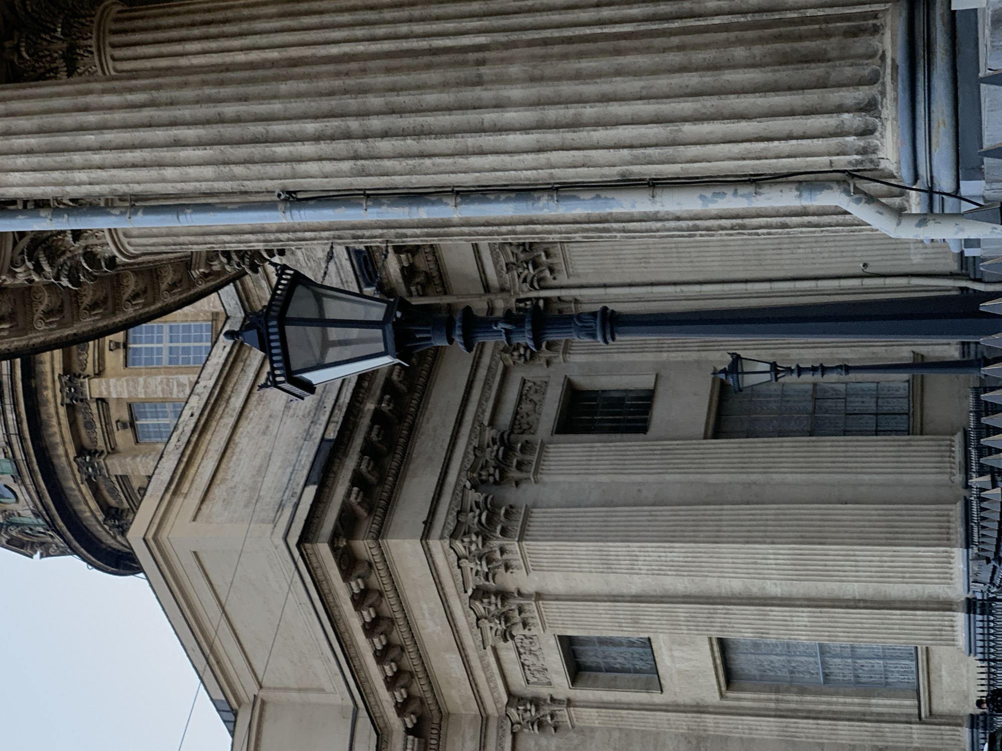 The image depicts an exterior view of an ornate building in Saint Petersburg, Russia. The architectural style is classical, featuring fluted columns, decorative moldings, and window openings. A black, ornate lamppost is affixed to the building's exterior. A section of fencing is visible on the right side of the image. Above the lamppost, a pipe runs along the top of the building. The sky is visible in the lower-left corner, suggesting a daytime setting. There are no people visible in the image.