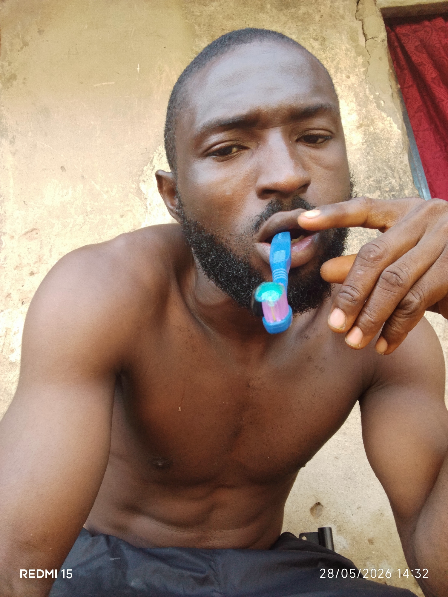 A shirtless man with dark skin, a beard, and a shaved head is holding a blue and pink toothbrush in his mouth. His right hand is raised, and one finger is extended. The background consists of a beige wall with some discoloration, and a red curtain is visible in the upper right corner. The photo was taken with a Redmi 15 on May 28, 2026, at 14:32. The image metadata does not confirm a location in Sabon Tasha, Nigeria.