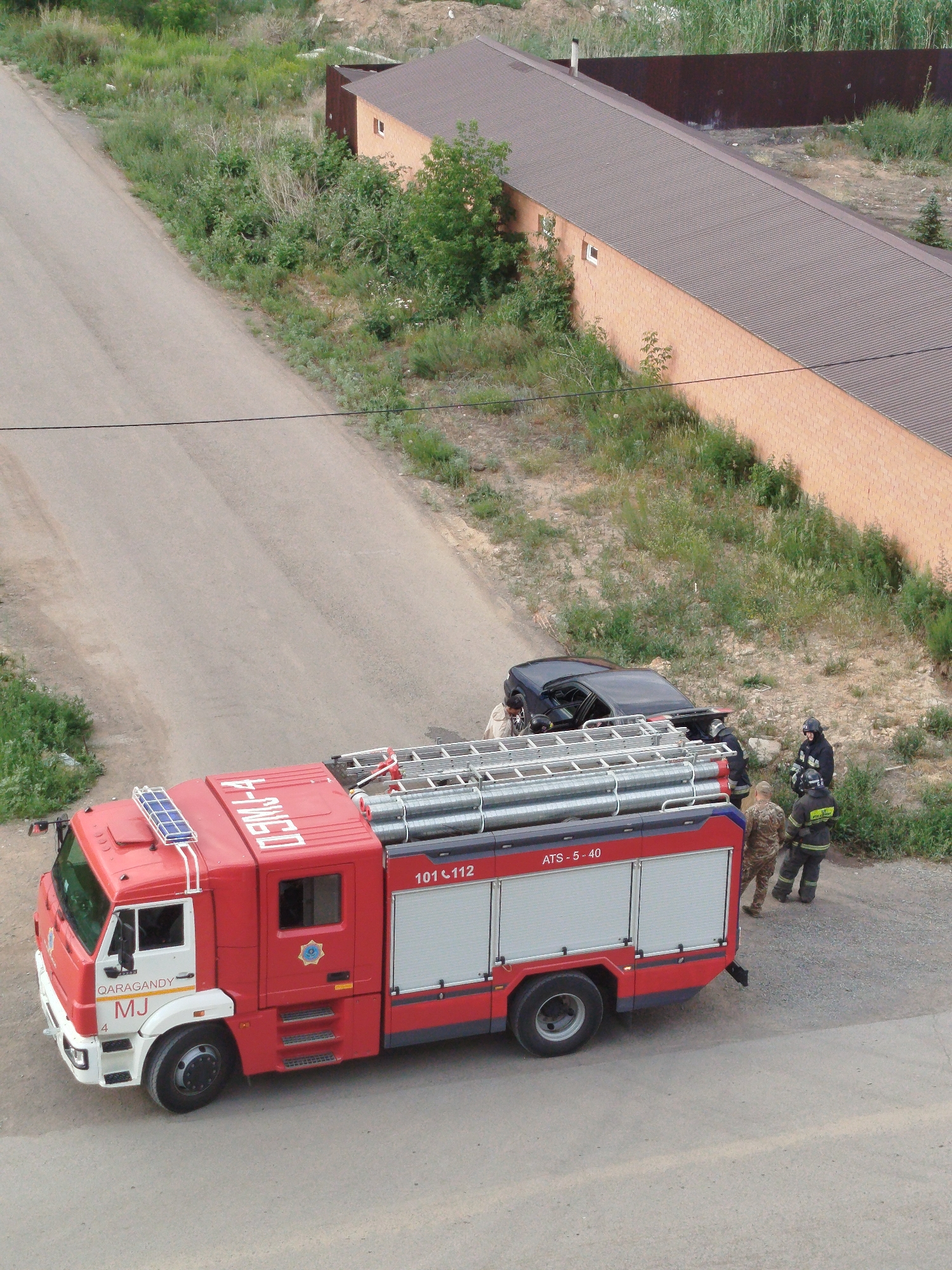 An aerial, high-angle medium shot captures a fire truck and a black car on a paved road adjacent to a grassy embankment and an orange brick building with a brown roof. The fire truck, a red vehicle with white accents, is parked on the road, its side facing the camera. The truck has "ATS - 5 - 40" and "101 112" visible on its side. A blue solar panel is mounted on the roof of the cab. A black car is positioned on the grassy embankment, appearing to be at an angle. Several individuals, dressed in what appear to be firefighter uniforms, are gathered near the black car and the fire truck. One person in a camouflage uniform is standing to the left of two individuals in dark firefighter gear. The environment suggests a potentially active scene, possibly a roadside incident or training exercise. The city of Qarağandı city administration, Kazakhstan is mentioned as context for the location.