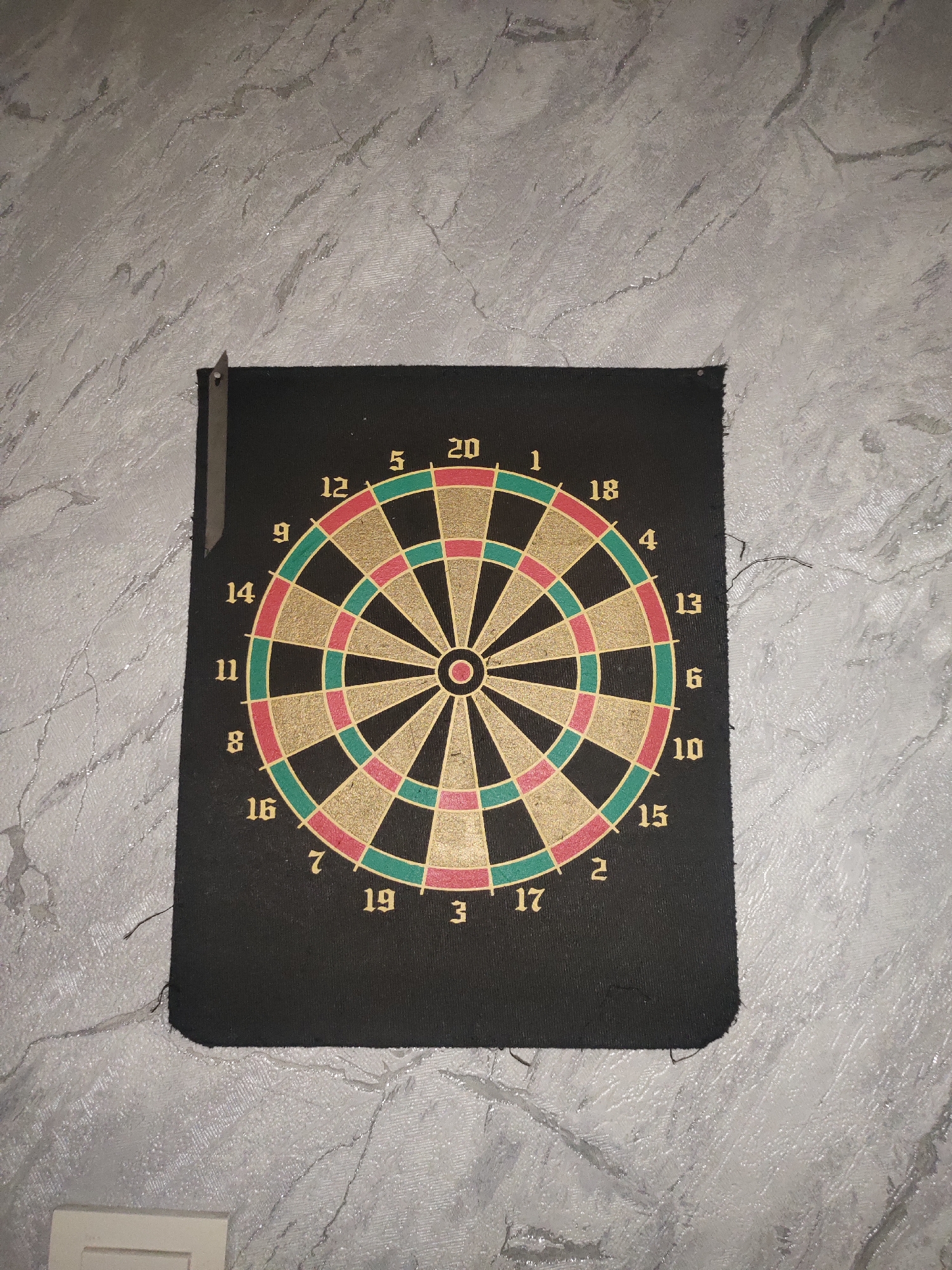 A dartboard is mounted on a textured grey wall. The dartboard is circular with numbered sections. The numbers visible are 20, 5, 12, 9, 14, 11, 8, 16, 7, 19, 3, 17, 2, 15, 10, 6, 13, 4, 18, and 1. The dartboard is black with colored segments in red, green, and gold. There is a small metal object attached to the top left corner of the dartboard. The wall appears to be a roughly textured plaster. The scene is indoors. There are no people or animals present. The image does not contain any information that would allow for determining its location in the city of Esik, Kazakhstan.