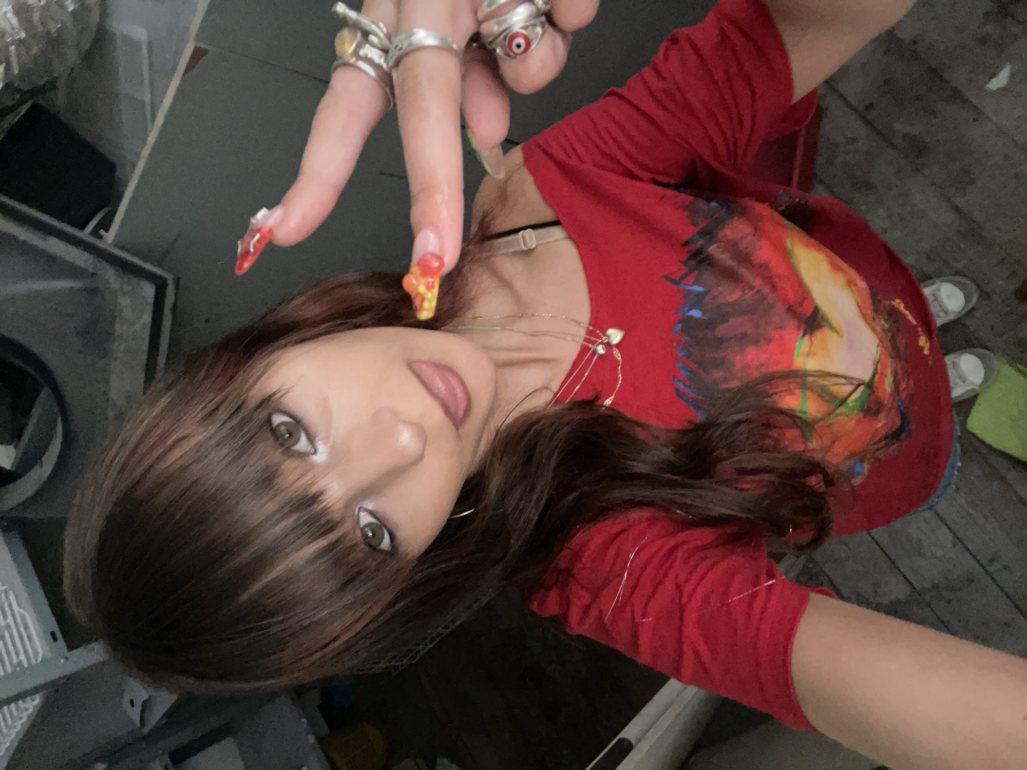 The image shows a young woman with brown hair and bangs, wearing a red off-the-shoulder shirt with a graphic print. She is sticking her tongue out and holding up a finger with a pizza-slice-shaped nail art. She is wearing several rings on her fingers. The background appears to be an interior space with industrial-looking equipment and wooden flooring. There is no indication that this image was taken in Raanana, Israel.