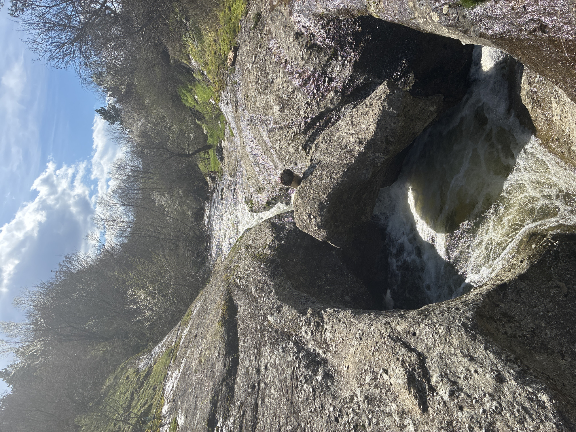 The image depicts a cascade of water flowing through a rocky terrain, possibly part of a river or stream. The scene is dominated by large, gray rocks with visible textures and crevices. The water is turbulent and foamy as it rushes through the rocks, creating a small waterfall or rapid. The surrounding environment includes vegetation, specifically trees and some patches of green moss on the rocks. The sky is visible in the upper left corner, displaying a clear blue color with white clouds. Based on the prompt, the location is Brezen, Bulgaria.