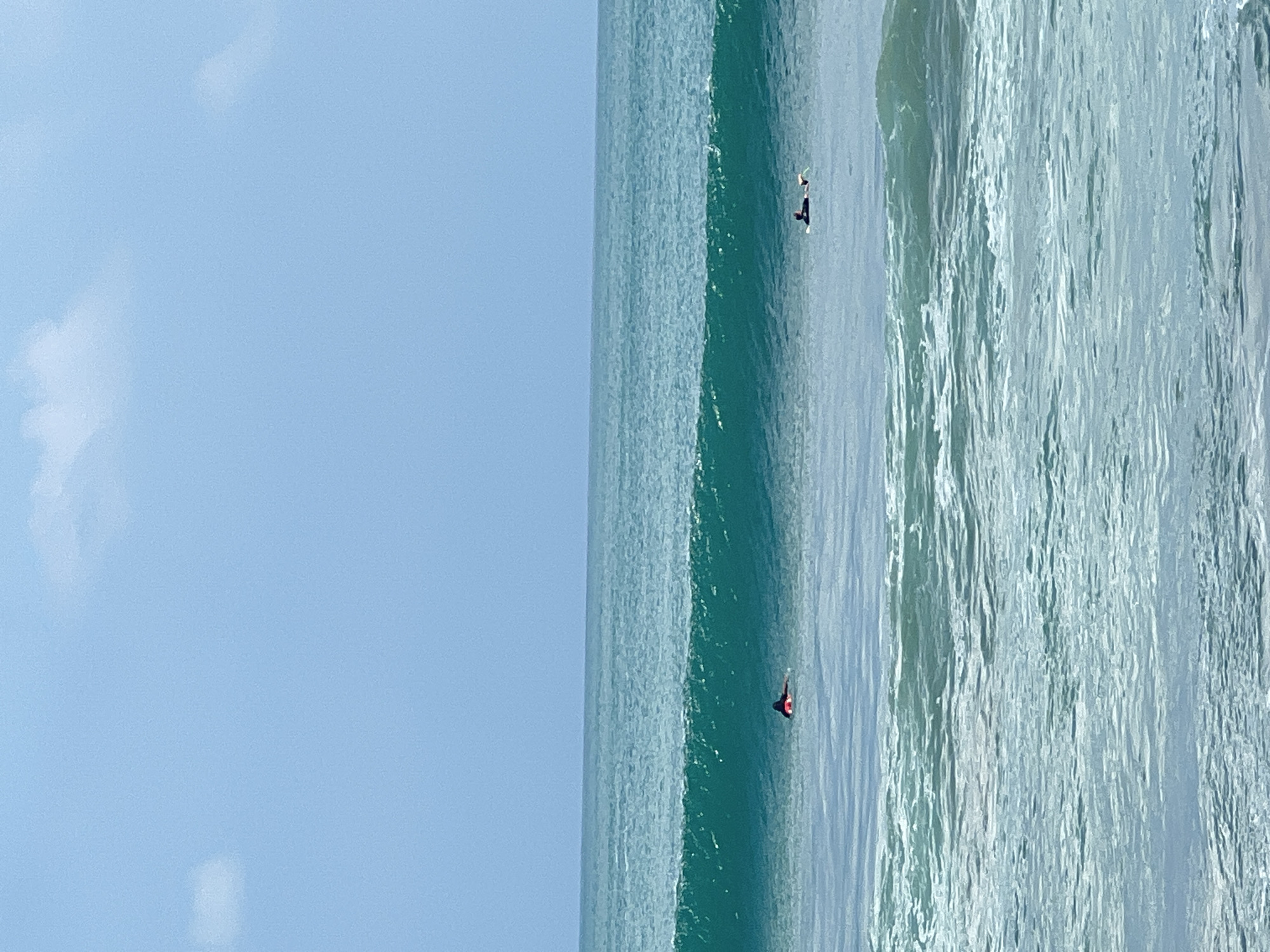 The image depicts a tranquil ocean scene in Kuta, Indonesia. The sky is a clear, light blue with a few wispy clouds scattered across the horizon. The ocean is a vibrant turquoise, with a large wave forming in the middle ground, its cresting white foam indicating its power and movement. Two surfers are visible in the water. One, wearing a red top, is lying on their surfboard closer to the breaking wave. Further out, another surfer, dressed in black, is also lying on their board, seemingly waiting for a wave. The foreground shows the gentle undulation of smaller waves with foamy white caps, suggesting a calm, yet dynamic, marine environment. The overall impression is one of peaceful coexistence with nature, with the surfers engaging in their sport against the backdrop of the vast ocean and serene sky.