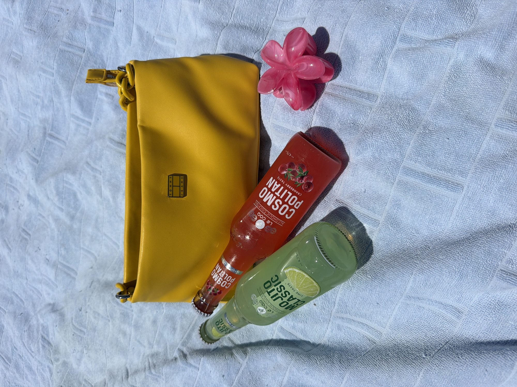 The image shows a yellow Tommy Jeans bag, a pink flower-shaped hair clip, a bottle of Cosmo Politian alcoholic cocktail, and a bottle of Mojito Classic alcoholic cocktail, all lying on a blue towel. The photograph appears to be taken in bright sunlight. There is no information to suggest the location is Rafailovići, Montenegro.