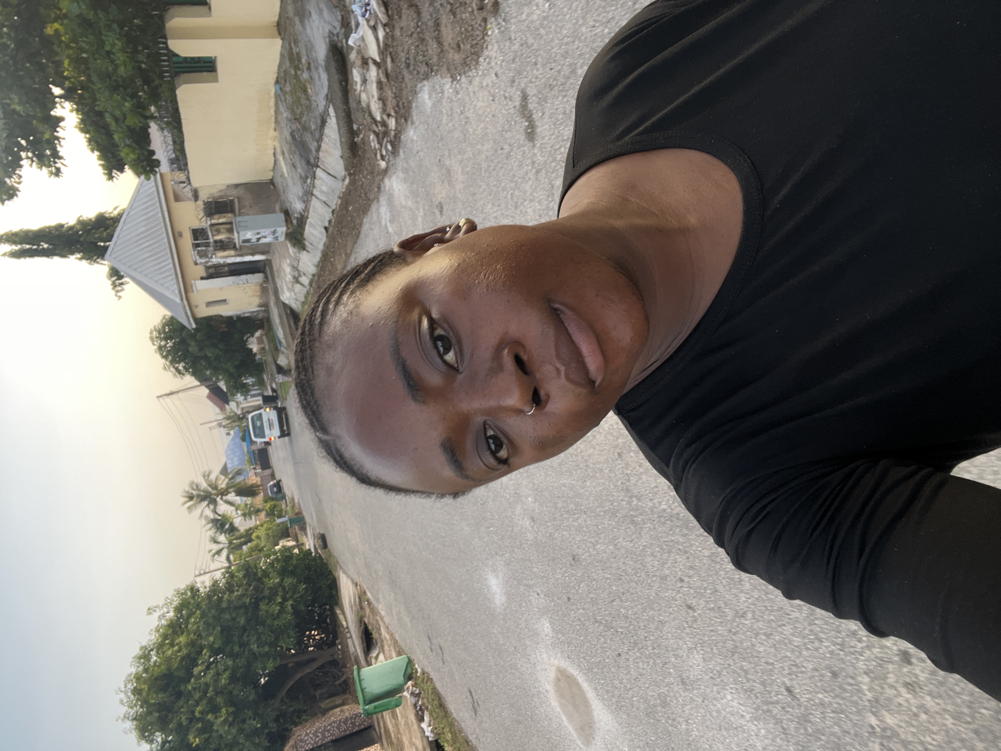 An outdoor selfie captures a person on a street in what appears to be an urban or suburban area of Abuja, Nigeria. The individual, positioned prominently in the foreground, has dark skin, wears a plain black long-sleeved top, and has braided hair. They are looking directly at the camera with a neutral to slightly pleasant expression and wear a small nose ring.
The background reveals a street scene under a bright, hazy sky, suggesting either early morning or late afternoon. To the left, a light-colored building with a grey corrugated metal roof is visible, partially obscured by green trees and foliage. The ground near the building consists of dirt and some paved sections.
The main thoroughfare is an asphalt road, extending into the mid-ground where a white vehicle, resembling a van or small bus, is driving. More buildings, utility poles, and scattered greenery, including palm trees, line the distant parts of the road. A green trash can is visible on the side of the road in the lower left portion of the image. There is no visible text in the media file.