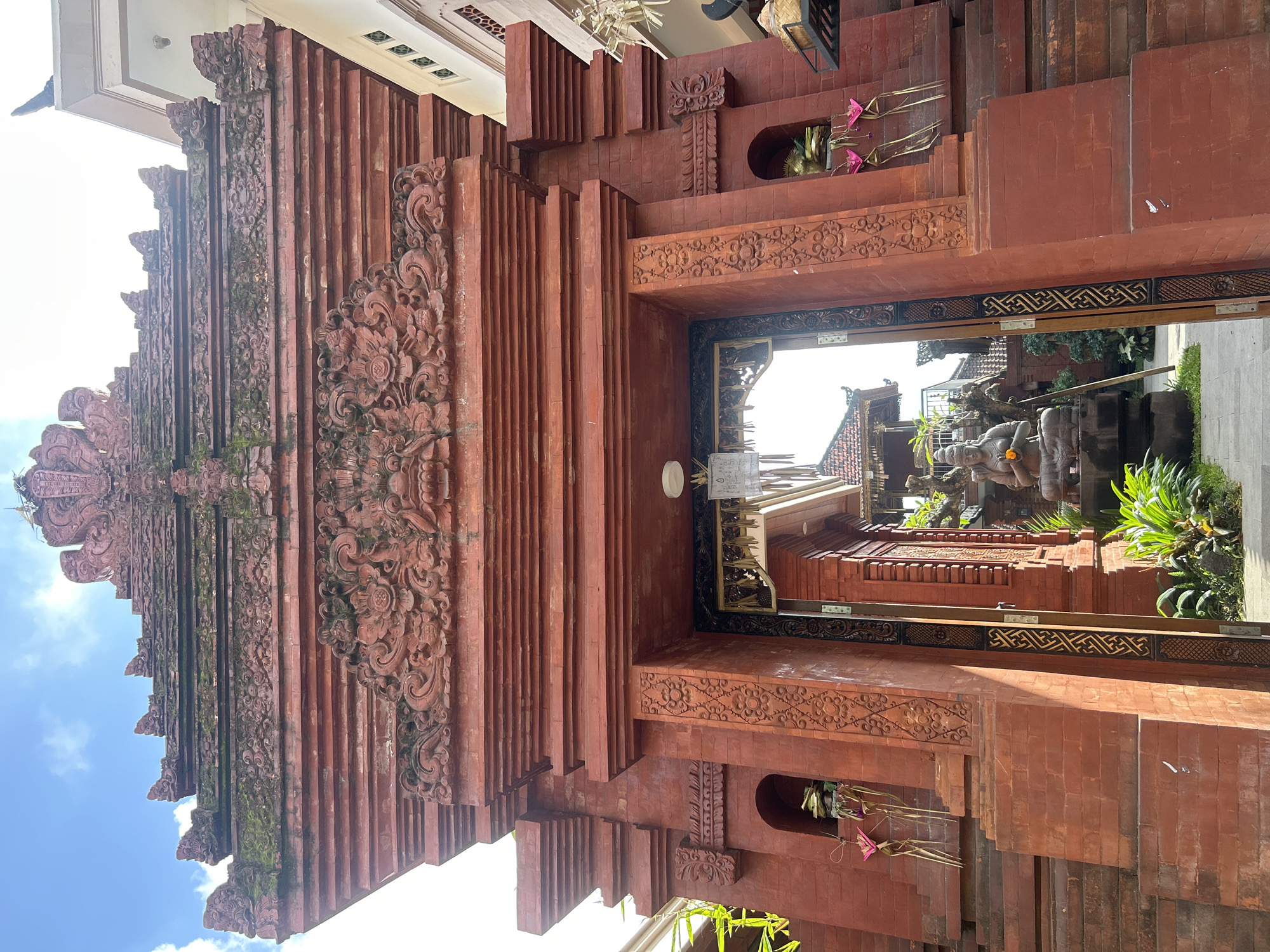 The image shows an ornate red brick gateway in North Kuta, Indonesia. The gateway is adorned with intricate carvings and topped with a tiered structure, parts of which are covered in moss. On either side of the entrance, small alcoves contain offerings, including pink flowers and what appear to be palm fronds. The gateway opens into a courtyard where a stone statue of a seated figure, possibly Buddha, is visible, holding an orange flower. Lush green plants surround the statue and extend to the base of the gateway. The sky above is blue with scattered white clouds. The overall scene suggests a place of spiritual or cultural significance.