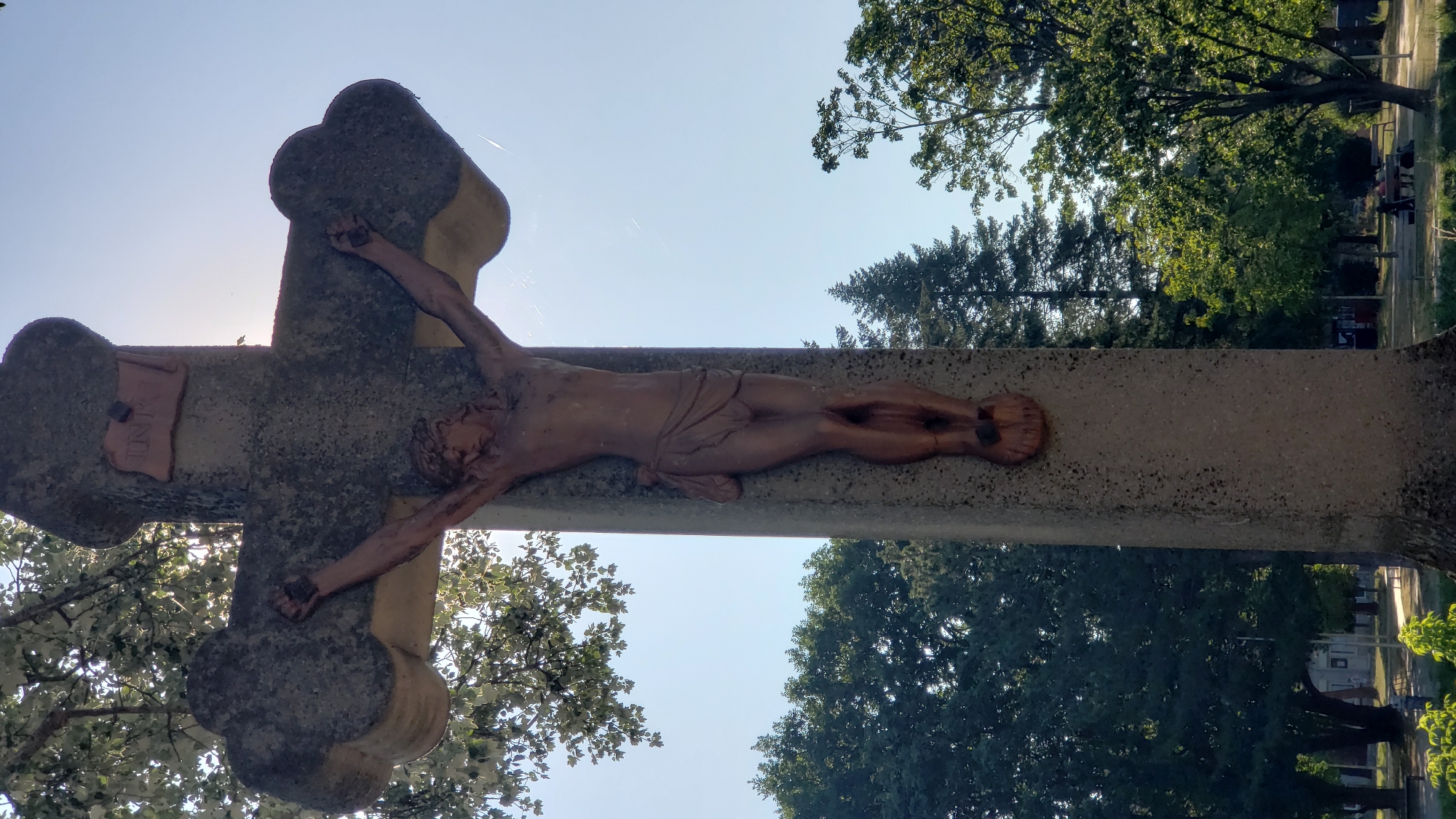 A stone crucifix stands in a park, possibly in Prešov, Slovakia, with a depiction of Jesus Christ. The crucifix is weathered, with the figure of Jesus appearing to be made of a different material, possibly painted wood or resin. The inscription "INRI" is visible at the top of the cross. The background features lush green trees, a clear blue sky, and a path with benches where a few people are seated. The sun is shining, casting light through the trees and creating a halo effect around the crucifix.