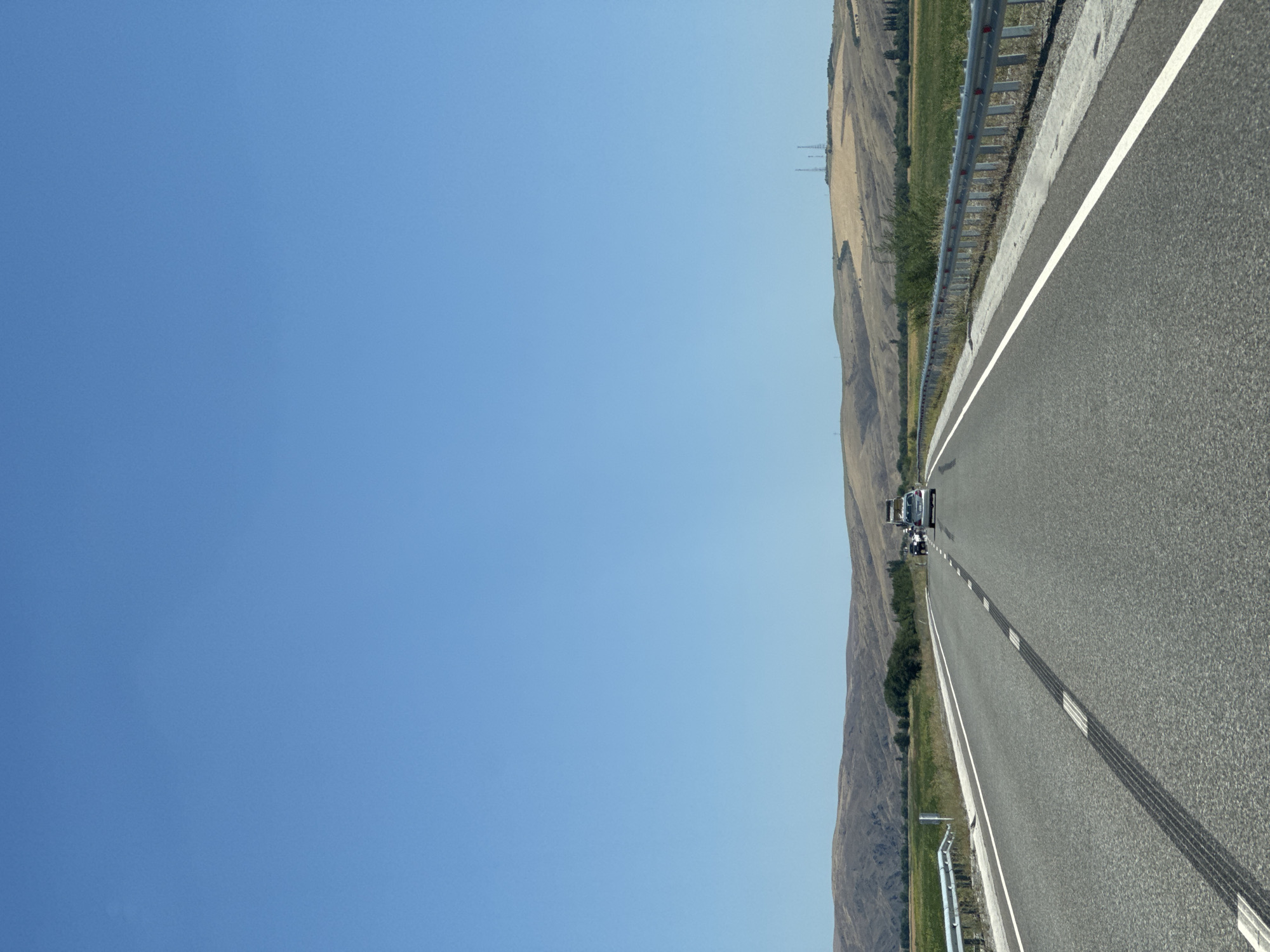 The image shows a clear, bright, daytime scene on a highway. The sky is a solid, vibrant blue, taking up the majority of the frame. Below the sky, rolling hills dominate the horizon, their slopes a mix of dry, golden-brown grass and patches of green vegetation. There are at least three tall, thin radio towers visible on the highest peak of the hills in the background on the right side.
The highway itself is asphalt, with white lane markings. On the left side of the road, a metal guardrail runs along the shoulder, separated by a concrete barrier. On the right side of the road, there is also a metal guardrail. In the foreground, there are prominent tire marks on the asphalt, indicating recent braking or acceleration.
Several vehicles are present on the highway. In the middle distance, a silver SUV and a white car are driving away from the camera. Further back, other vehicles are visible, suggesting moderate traffic. Green fields are visible alongside the highway to the left, interspersed with trees. The overall impression is of a rural or semi-rural setting in Kazakhstan, possibly on a main road leading out of or into a populated area. The location provided, Qoilyq, Kazakhstan, aligns with the visual cues of a somewhat arid landscape with agricultural fields and highways.