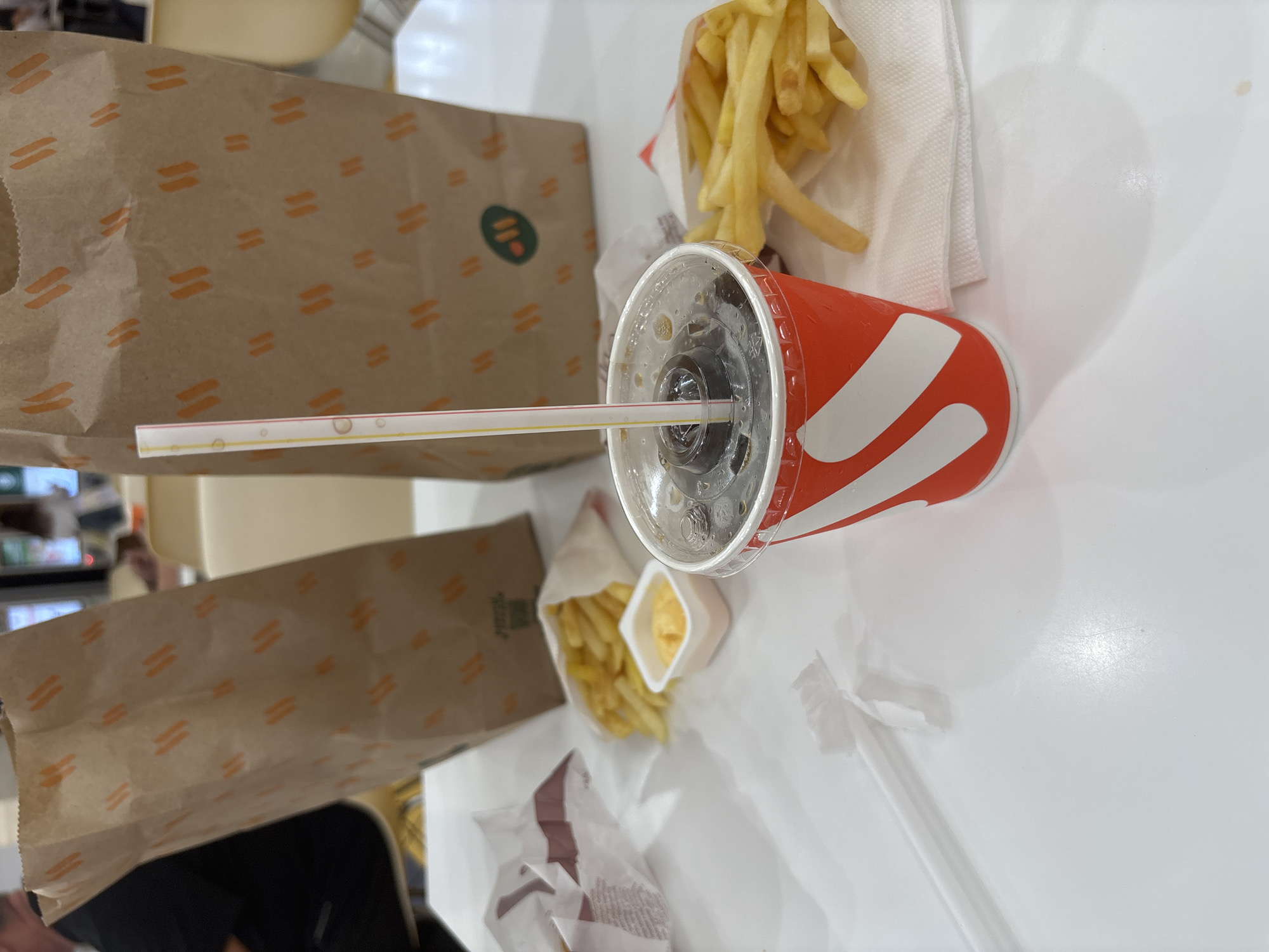 The image depicts a table at what appears to be a fast-food establishment, possibly in Kazan, Russia, given the context. In the foreground, a red and white paper cup with a clear plastic lid is filled with a dark beverage, likely soda, with ice cubes visible and a straw inserted. The cup has a distinctive red and white stripe pattern. Next to the cup, to the right, a portion of french fries is served on a white paper napkin. In the background, two brown paper bags with an orange patterned design are visible, suggesting takeaway food. One bag has a green circular logo with white text. Further to the left, another paper bag wrapper, white with red accents, is partially visible, along with a small white container holding an orange dipping sauce. French fries are also spilling out of a paper wrapper on the left side of the table. A person's shoulder and arm, dressed in a black shirt, are visible on the far left of the image, suggesting a dining companion or customer. The overall setting is a brightly lit indoor environment with a white tabletop.
