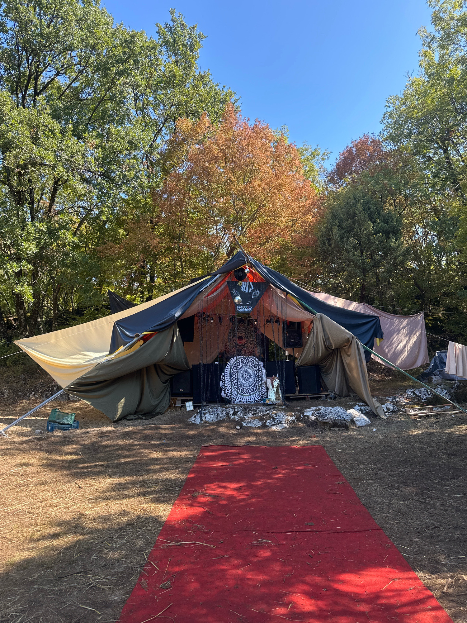 The image shows an outdoor setting with a large, multi-colored tent or canopy erected in a wooded area. The canopy is constructed from various fabrics, including tan, dark gray, and olive green, supported by poles. In front of the canopy, a red carpet is laid out on the ground, which is covered in dry grass and straw.
Behind the tent, there are trees with some leaves in autumn colors, suggesting a fall season. The sky is clear and blue. The tent appears to be set up for an event or gathering, with speakers visible inside and decorated with patterned cloths and what looks like some religious figurines. The overall scene suggests a bohemian or festival-like atmosphere.
This scene is not located in the city of Cetinje, Montenegro. It is an outdoor natural setting.