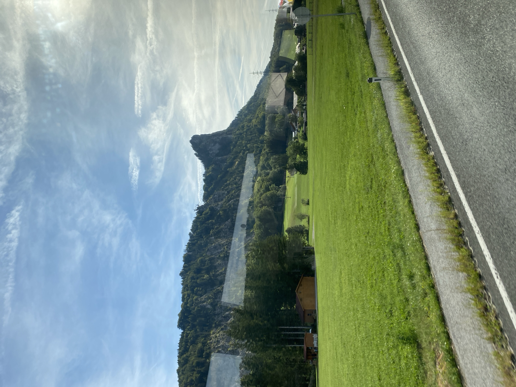 This is a scenic landscape photograph taken from a moving vehicle, likely a car or train, looking out at a rural Austrian scene. The foreground shows a paved road with a white line and a small white post marker on the right side. Adjacent to the road is a strip of grass and then a large, vibrant green meadow. In the mid-ground, there are several small wooden buildings, including a red-roofed structure and a larger, light brown barn-like building with an arched opening. A dense line of tall evergreen trees runs behind these buildings, separating the meadow from the next level of the landscape.
Dominating the background is a majestic, rocky mountain with a jagged peak that juts sharply into the sky. The mountain is covered in lush green trees and vegetation, with visible rock faces. The sky is a bright blue, filled with wispy white clouds that are illuminated by sunlight, suggesting it is a clear, sunny day. In the distance, the faint outlines of power lines and a few scattered houses can be seen, indicating a small village or settlement nestled at the base of the mountain. The overall impression is one of natural beauty and tranquility in a mountainous region. The image is taken at an angle, suggesting the viewer is traveling along a road that runs parallel to the meadow and the base of the mountain. The presence of reflections on the windows suggests the photo was taken through glass.