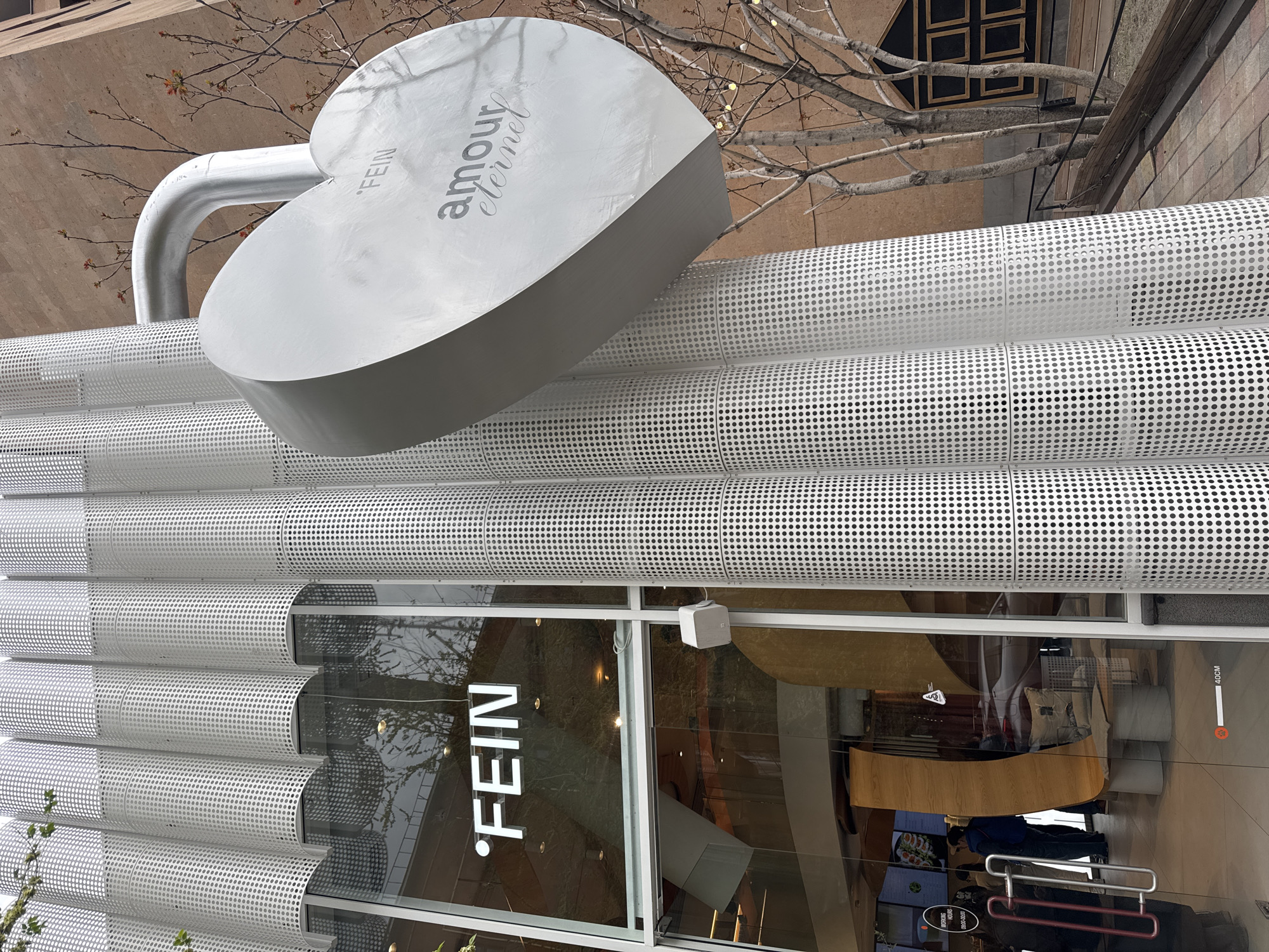 The image captures the facade of a modern commercial building in Yerevan, Armenia, under bright daylight. The building's exterior features a distinctive design with multiple horizontal rows of curved, perforated, silver-grey metal panels, creating a ribbed texture.
Prominently displayed above the ground-level windows is a large, metallic, heart-shaped sculpture. On its surface, the word "FEIN" is written vertically, and "amour éternel" is inscribed in a graceful cursive font. A curved, pipe-like structure extends from the left side of the heart, connecting to the building.
Below the heart sculpture, large glass windows span the ground floor. These windows reflect the surrounding environment, including bare tree branches with small green buds and fragments of nearby structures. Through the glass, parts of the building's interior are visible, featuring wooden and upholstered furnishings, suggesting a cafe or restaurant. The illuminated word "FEIN" is also displayed horizontally on one of the window panels. Further details visible in or through the glass include a "140CM" height marker and a sign indicating "OPENING HOURS 09:00-22:00". The scene depicts a contemporary urban setting with a focus on architectural detail and a symbolic heart motif.